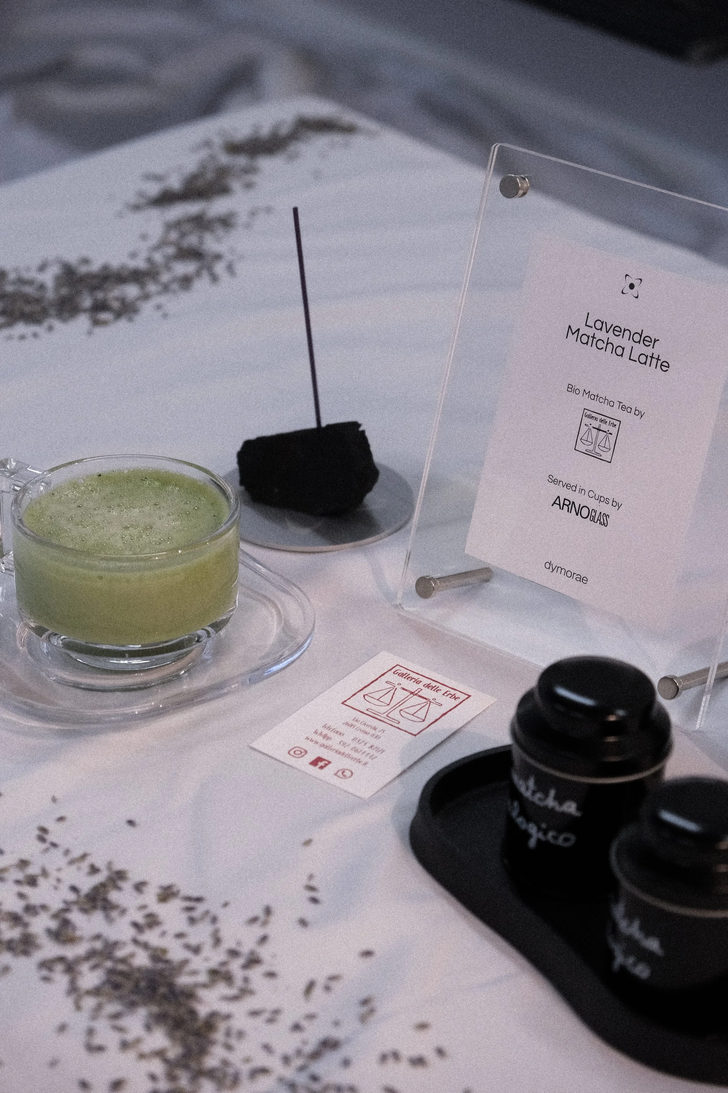 A small glass cup filled with a light green matcha latte on a clear glass saucer, with a spoon inside. Behind it, a black rock-like object with a thin stick or incense stick inserted. To the right, a white sign with black text reads "Lavender Matcha 