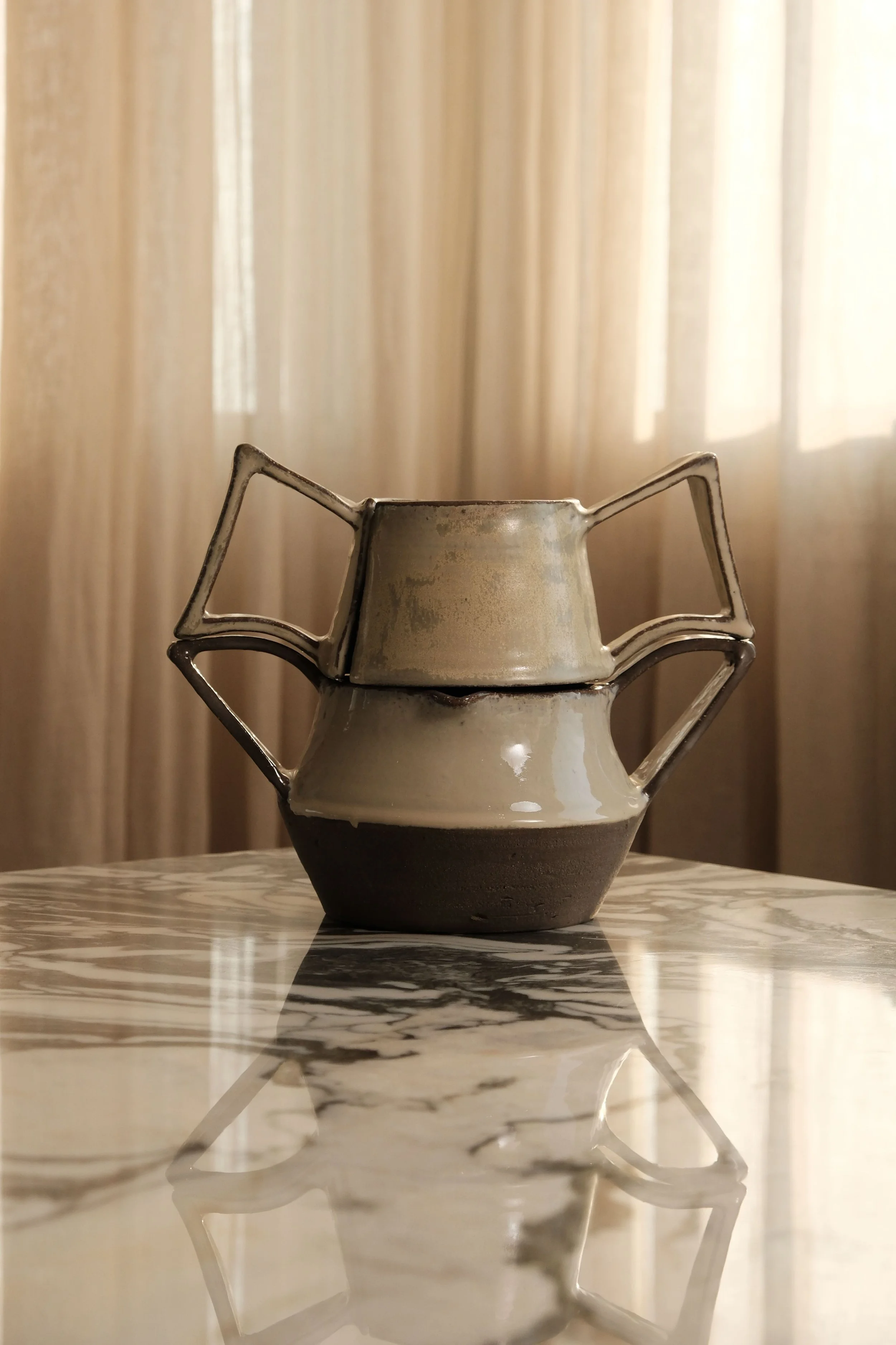 A ceramic vase with angular metal handles, placed on a reflective marble surface with a black and white pattern, in front of beige curtains.