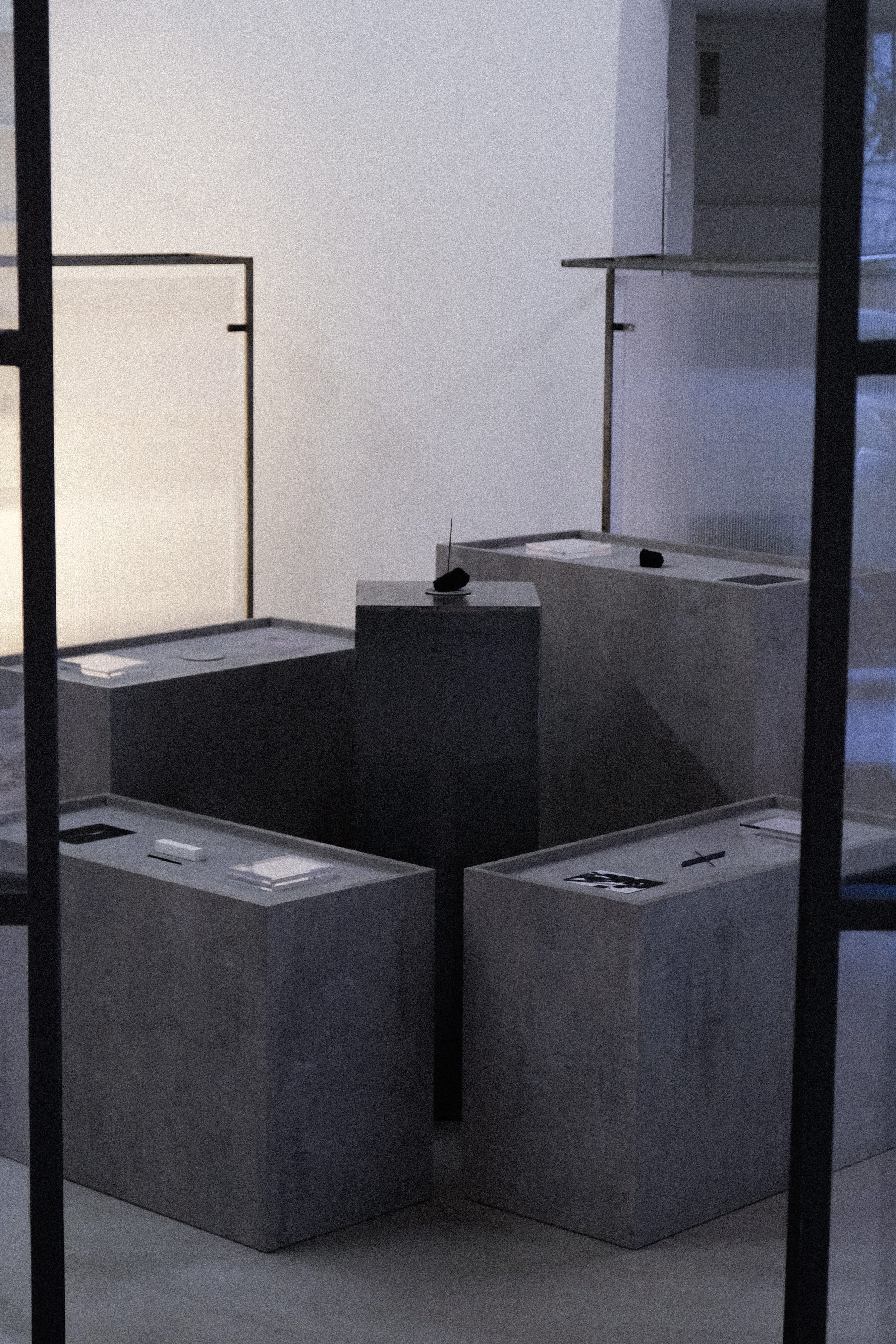 A minimalist art installation featuring several gray rectangular platforms with various printed images and objects on top. The setting appears to be an art gallery or exhibit space with a clean, modern aesthetic.