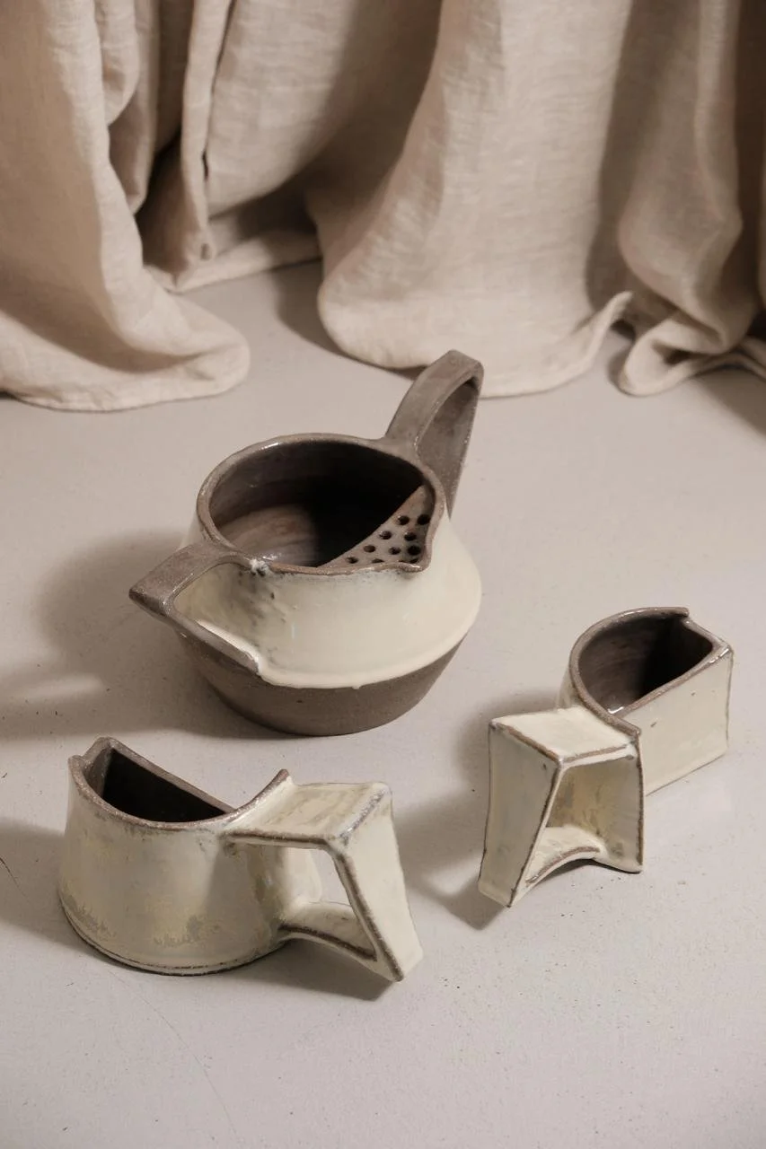 Set of three handmade ceramic cups with a beige glaze and dark interior, placed on a white surface with a light-colored fabric backdrop.