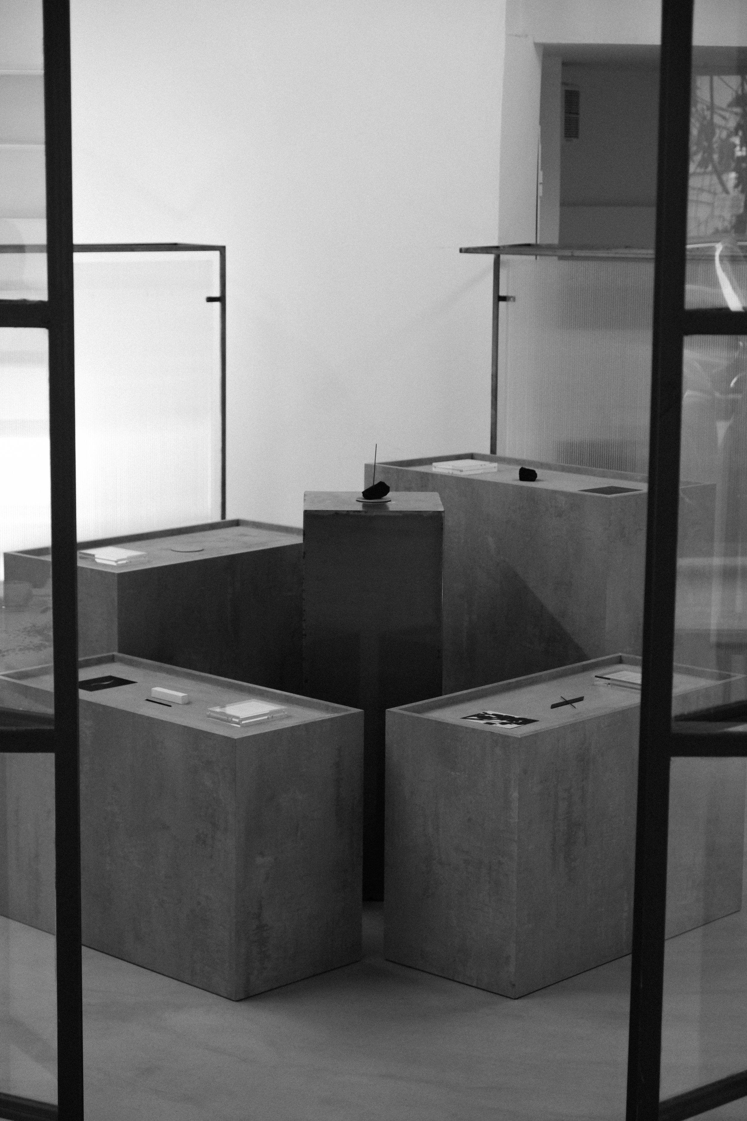 Exhibition display with four wooden pedestals holding papers and small objects, viewed through glass with black framing.