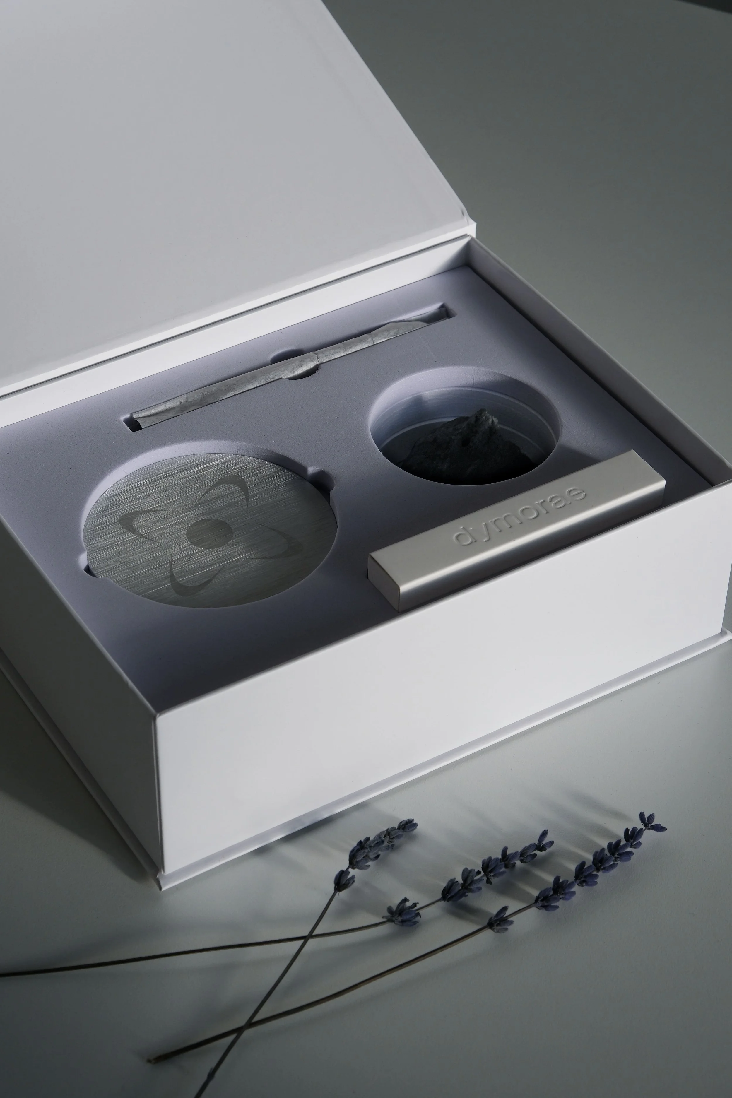 A white box with a silver and black device inside, labeled 'djmorae', and a small plastic trowel. Lavender sprigs are placed in front of the box.