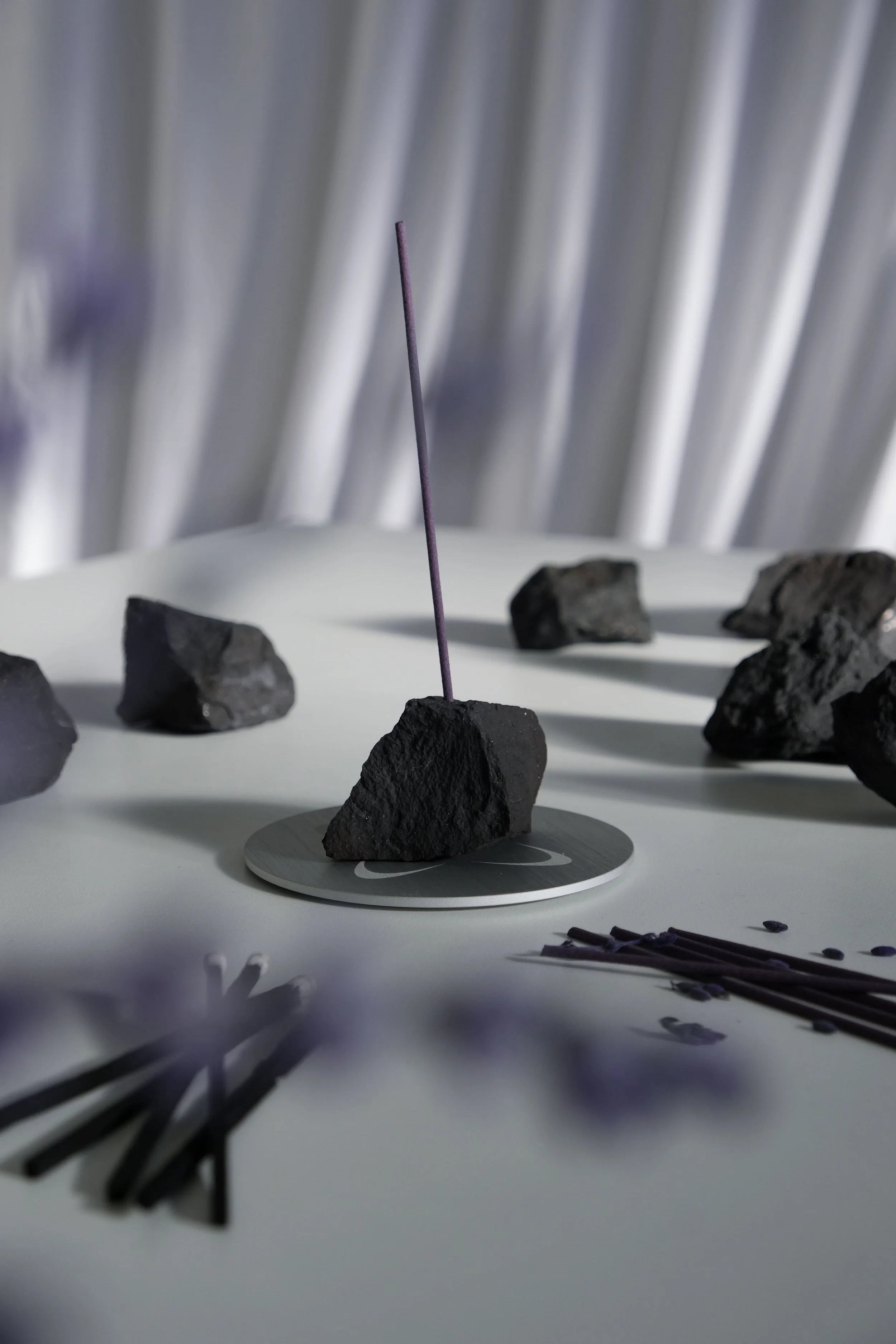 A collection of dark rocks on a white surface with some incense sticks and a circular stand, with a blurred background of metallic vertical lines.