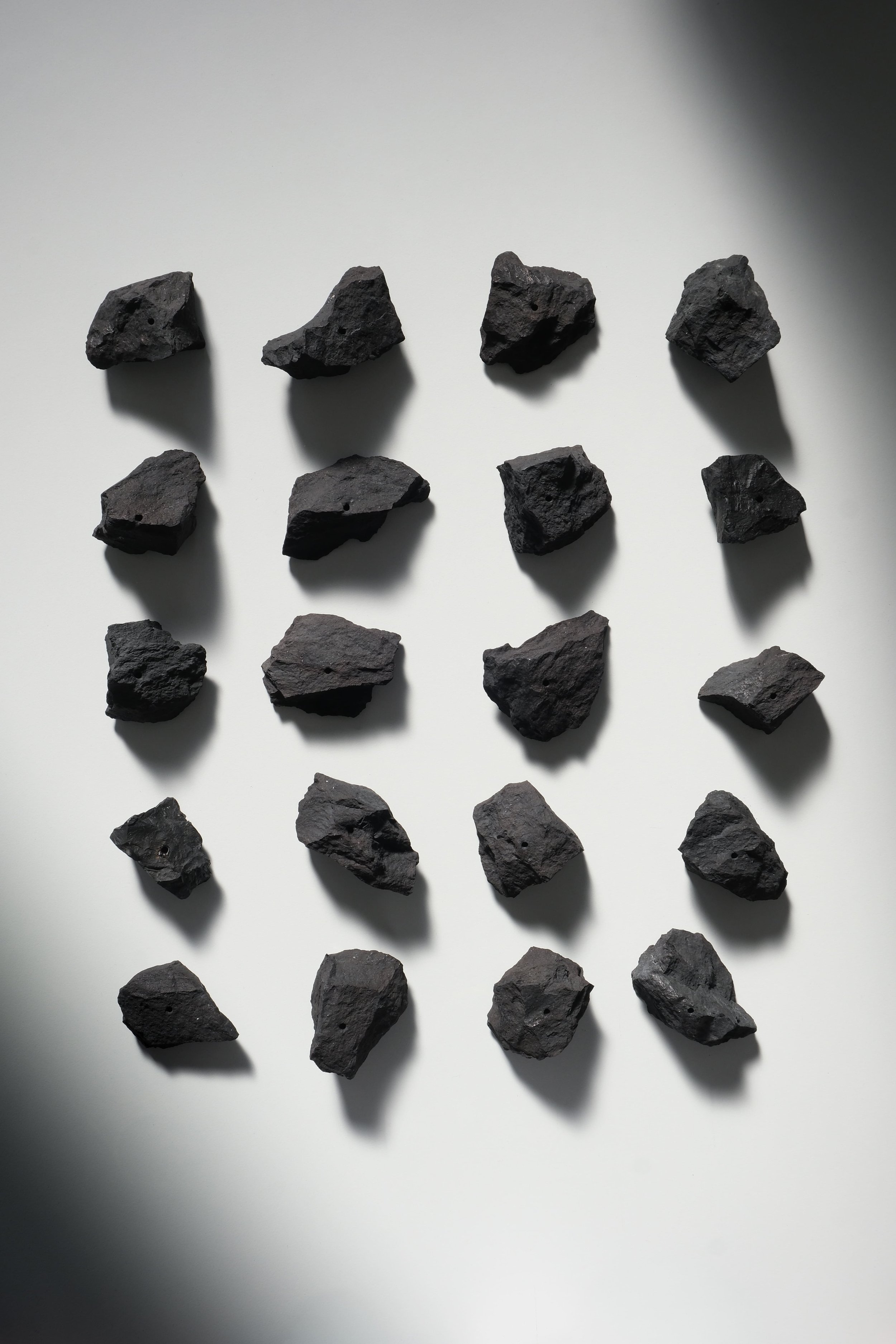 A flat surface with 15 black volcanic rocks arranged in a pattern, with shadows cast on the surface from a light source.