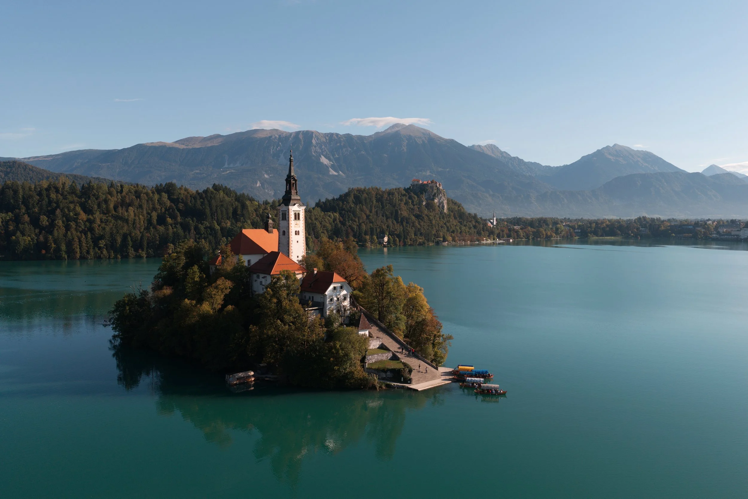 A small island with a church, trees, and buildings surrounded by a lake, with mountains in the background.