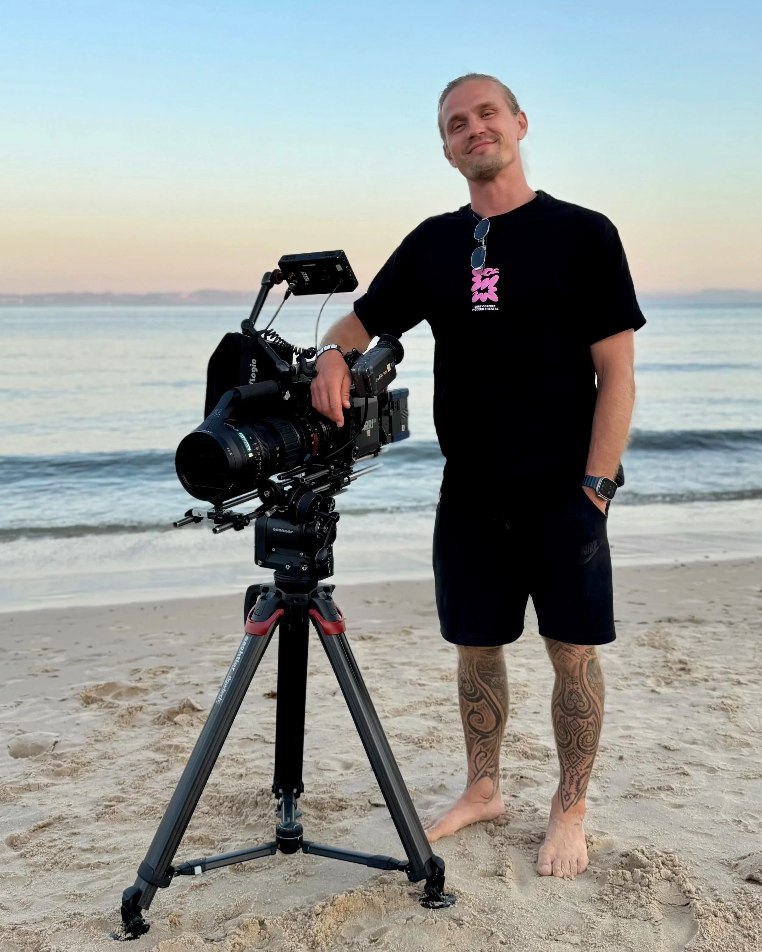 A man standing on a beach with a camera mounted on a tripod, wearing a black t-shirt, shorts, and a smartwatch, with tattoos on his legs, during sunset or sunrise.
