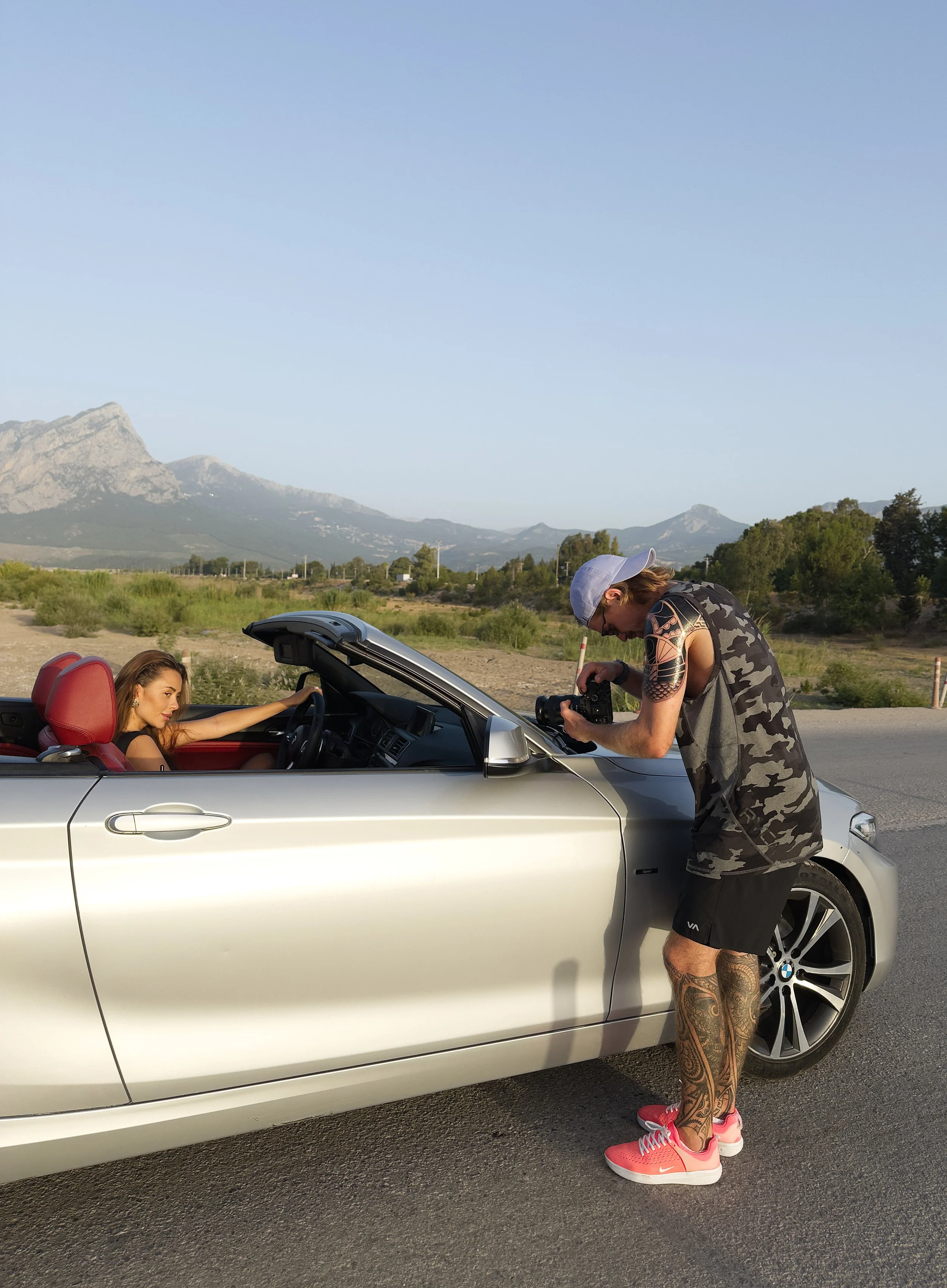 A woman sitting in a silver convertible car with red interior, being photographed by a man with tattoos on his legs, wearing a camouflage tank top, shorts, and sneakers, outdoors with mountains and greenery in the background.
