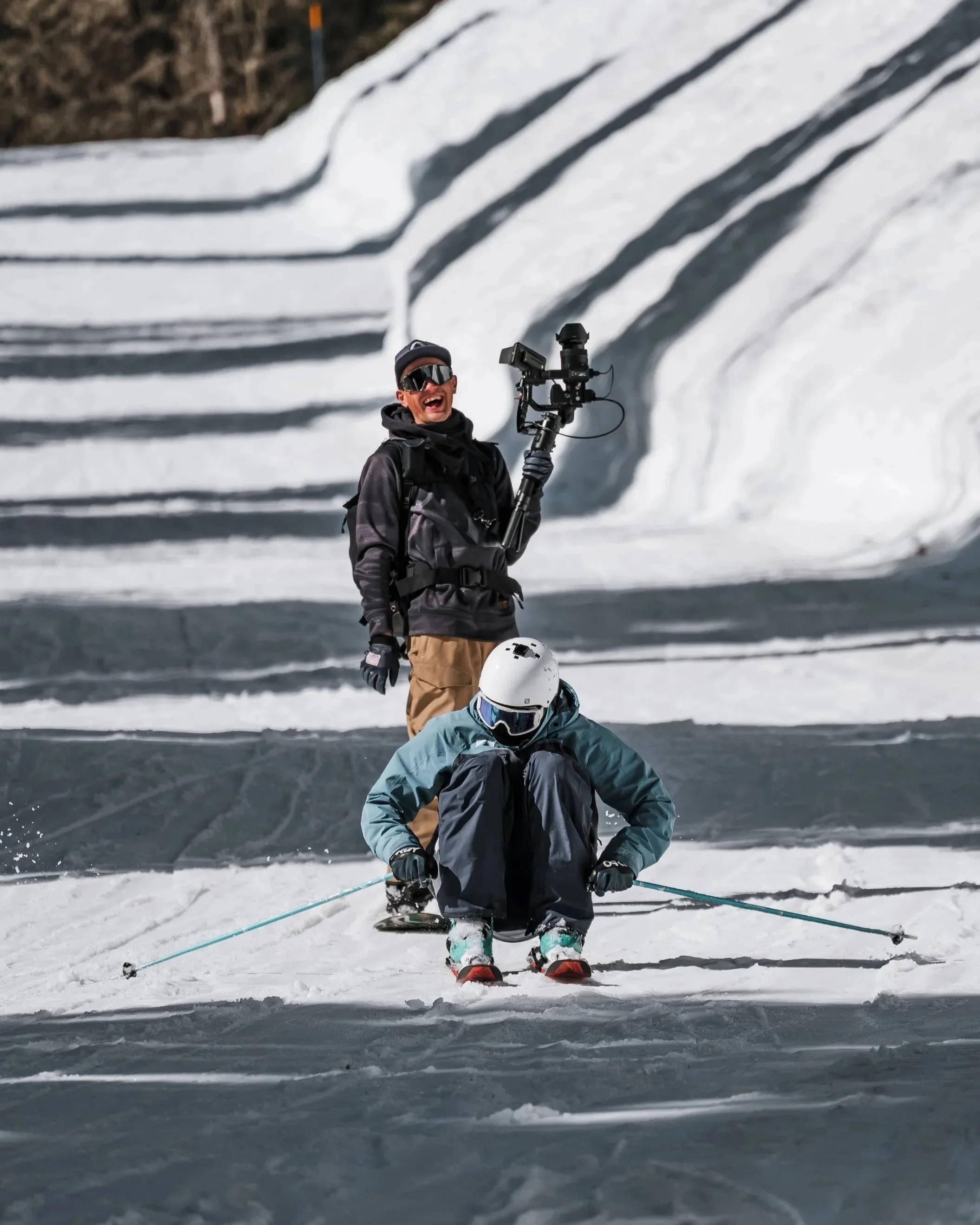 Two skiers, one standing and one kneeling, on snow-covered ground with mountainous background, and one person holding a professional video camera.
