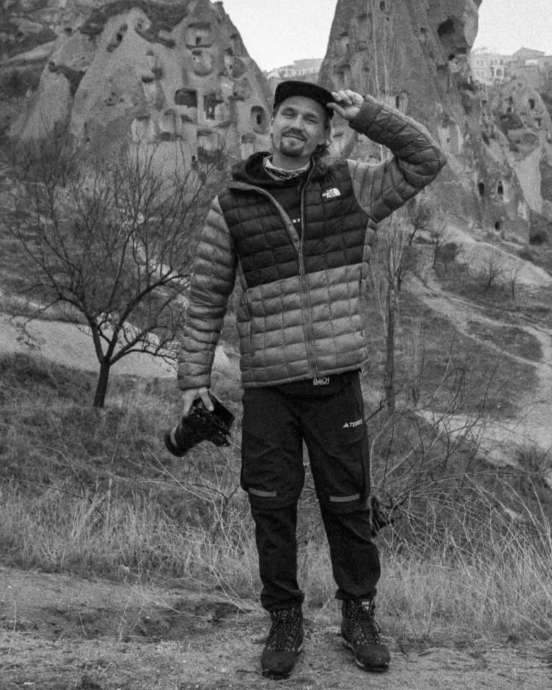 A man standing outdoors in a black and white photo, holding a camera in his left hand and adjusting his cap with his right hand, with unique rock formations and a few leafless trees in the background.