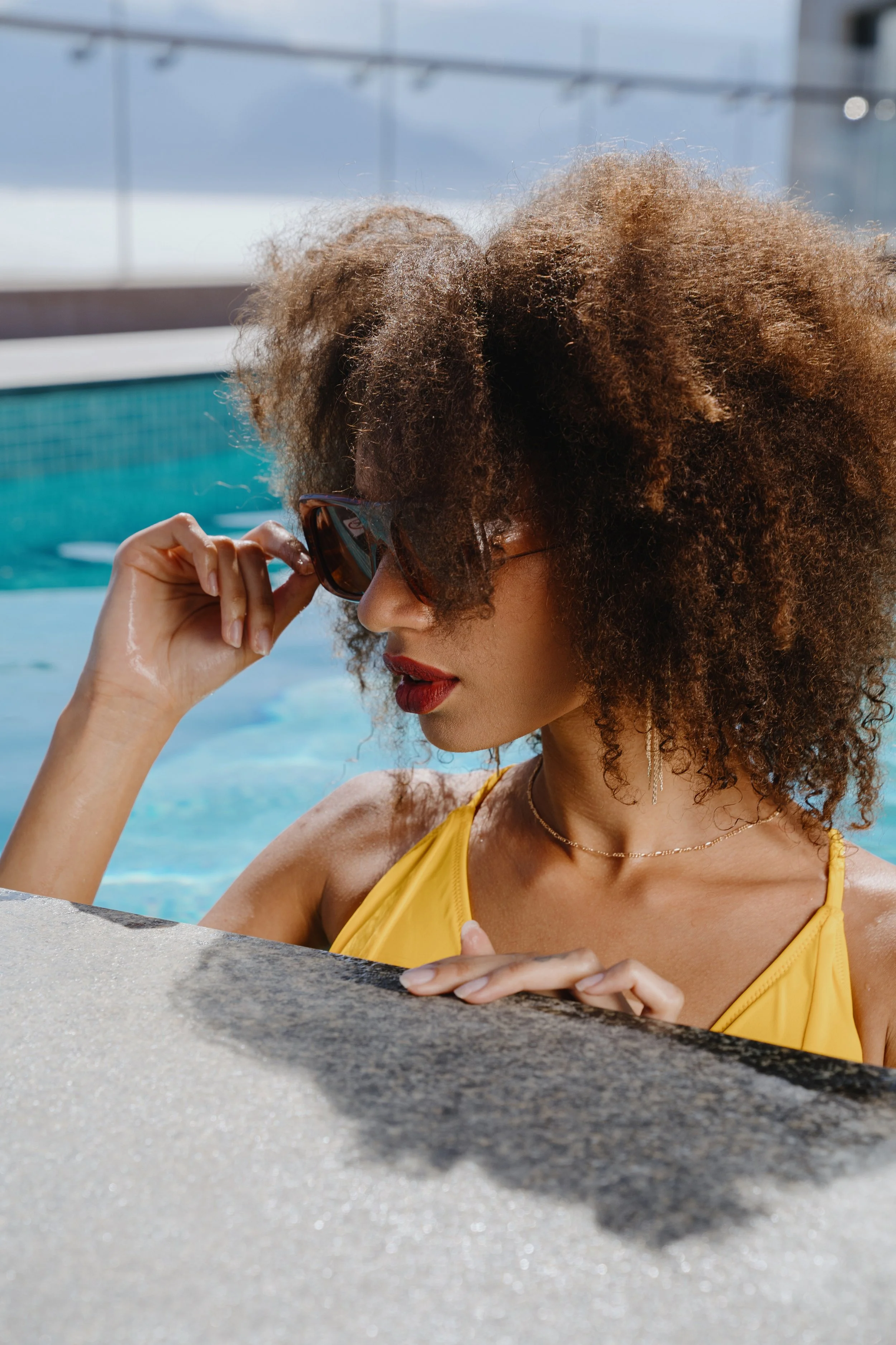 Woman with curly hair wearing sunglasses and a yellow swimsuit relaxing by a pool, holding her sunglasses with one hand.