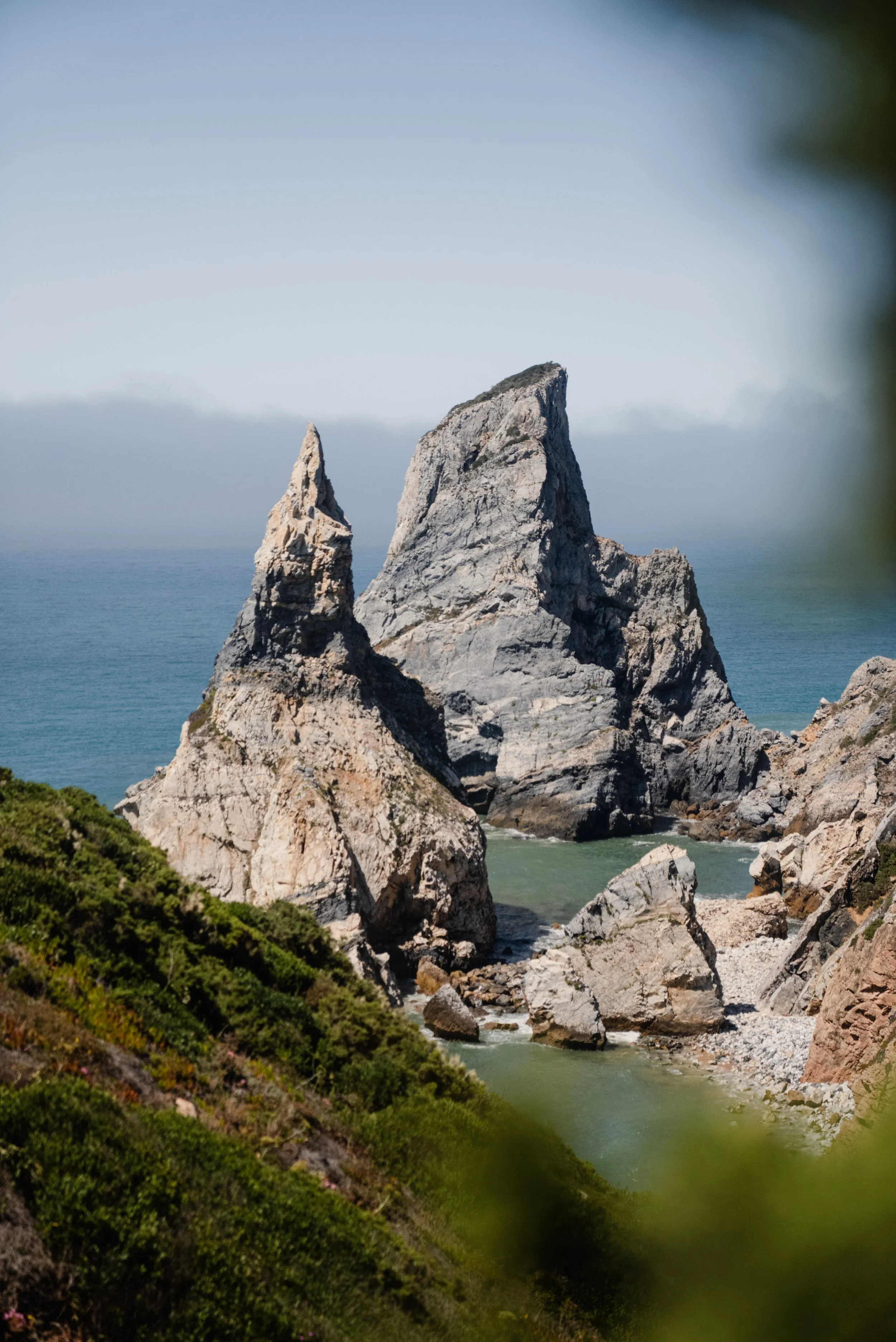 Three large rocky sea stacks rising from the ocean, with a green hillside in the foreground and a misty horizon.