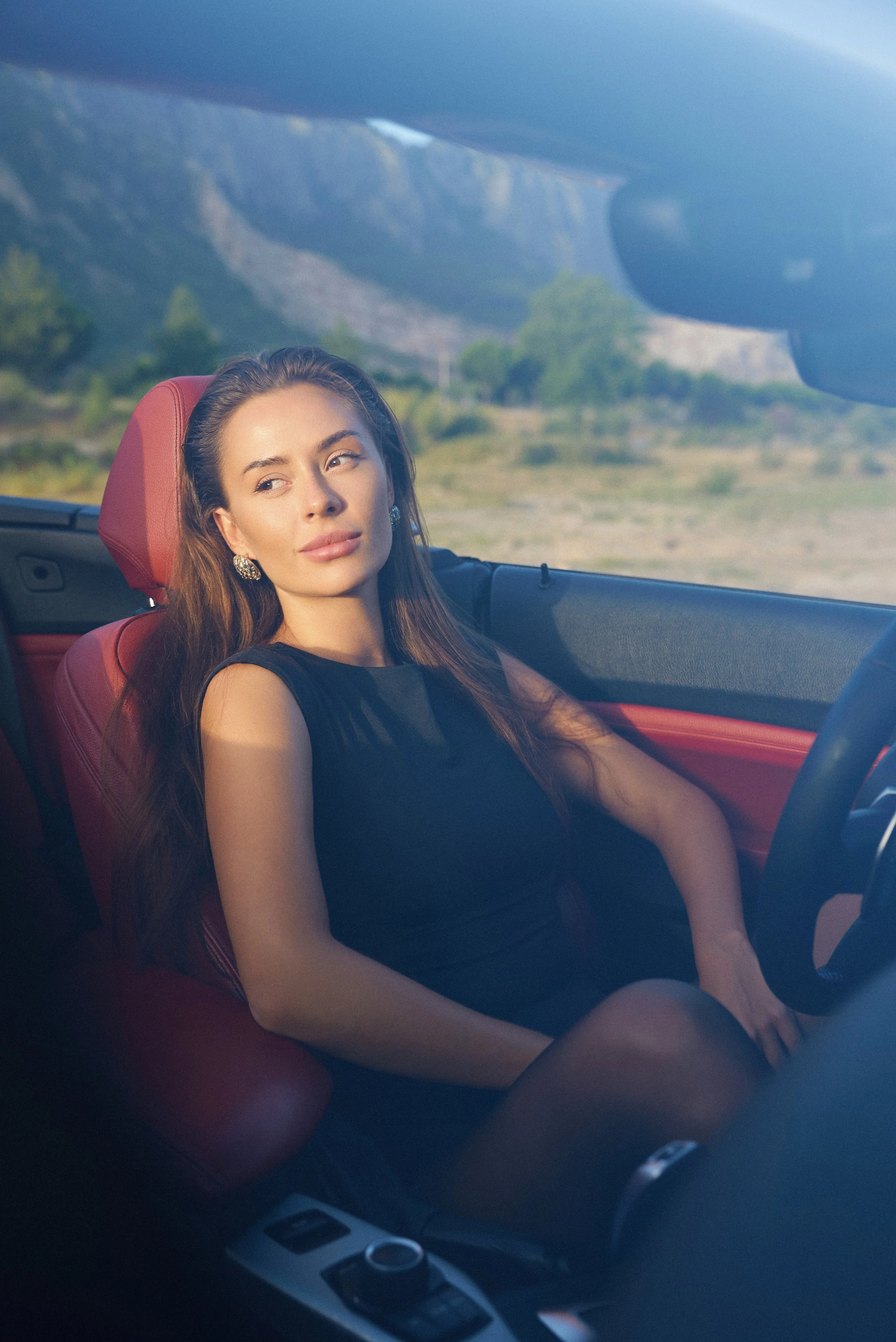 A woman with long brown hair and earrings sitting in the driver's seat of a car, looking out the window at a scenic landscape with mountains and trees.