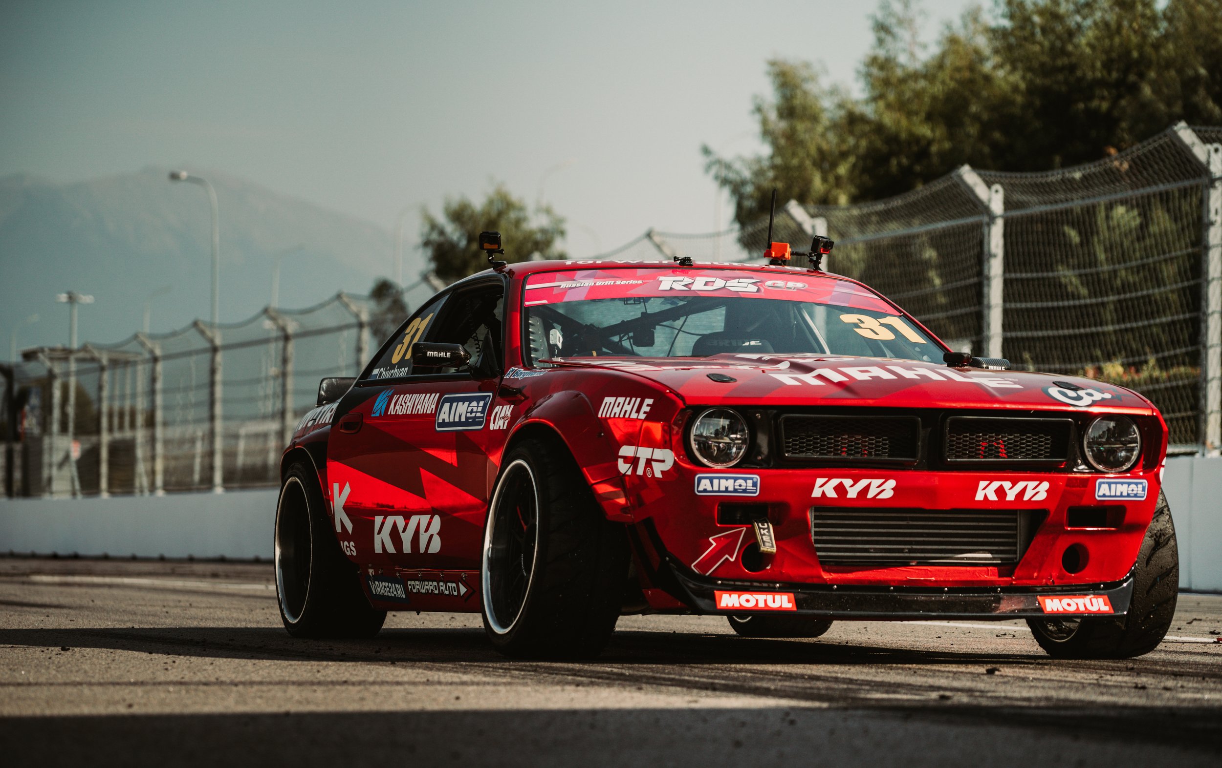 Red race car with various sponsor decals on a track, with a fence and trees in the background.