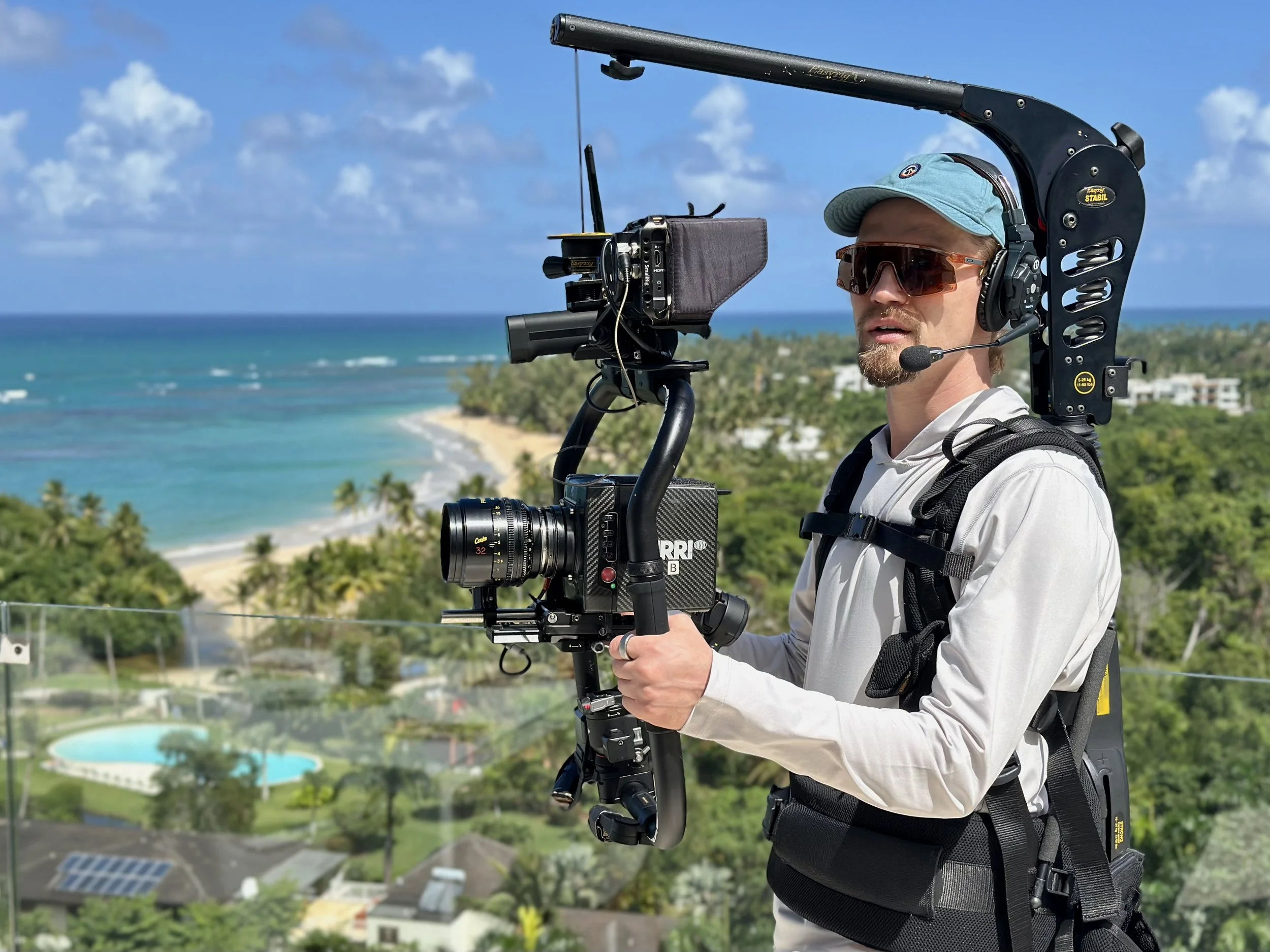 A man filming with a shoulder-mounted camera rig on a balcony overlooking a tropical beach and lush greenery.