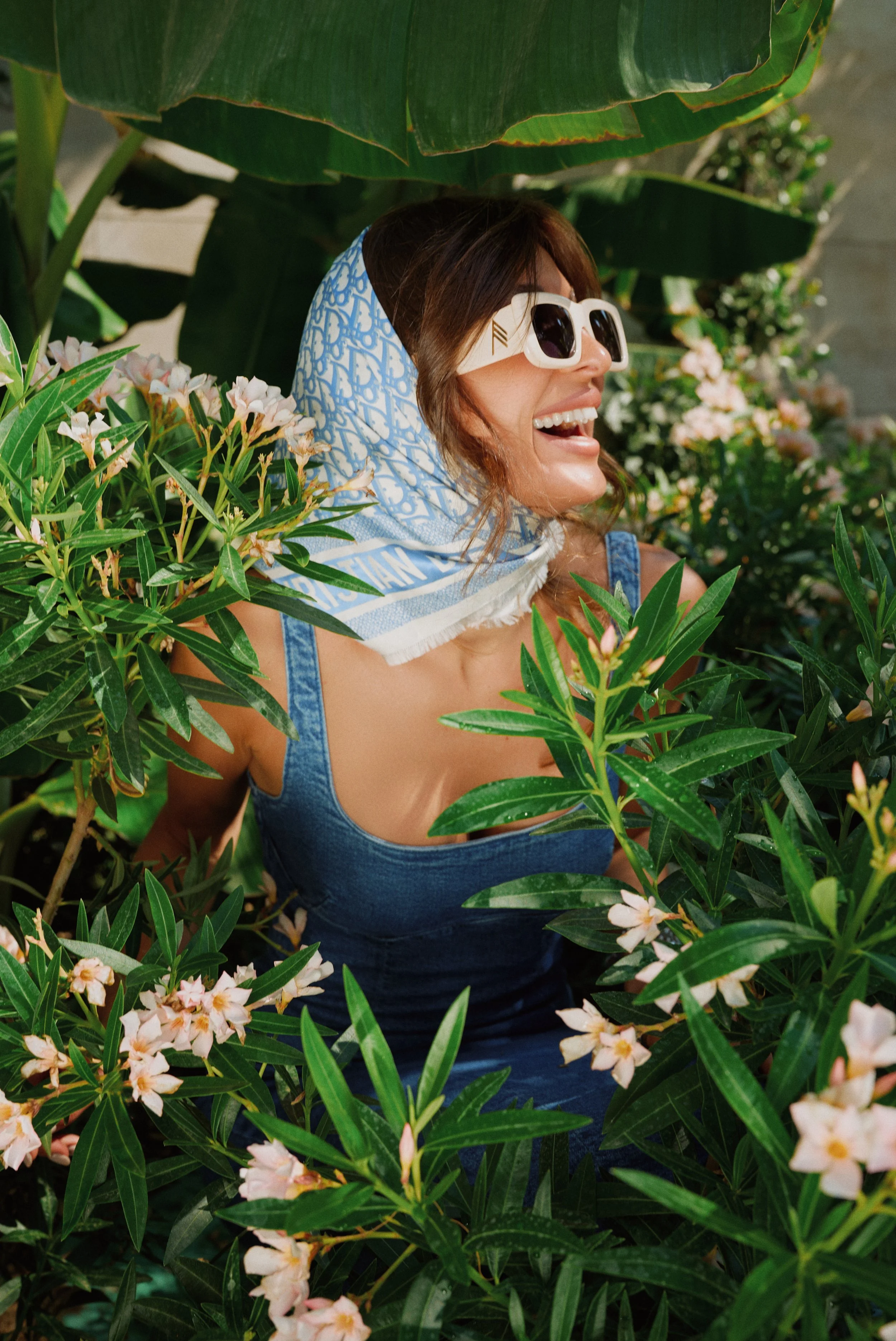 Woman wearing sunglasses and a blue scarf with white patterns, smiling and looking to the side, surrounded by lush green plants and pink flowers.