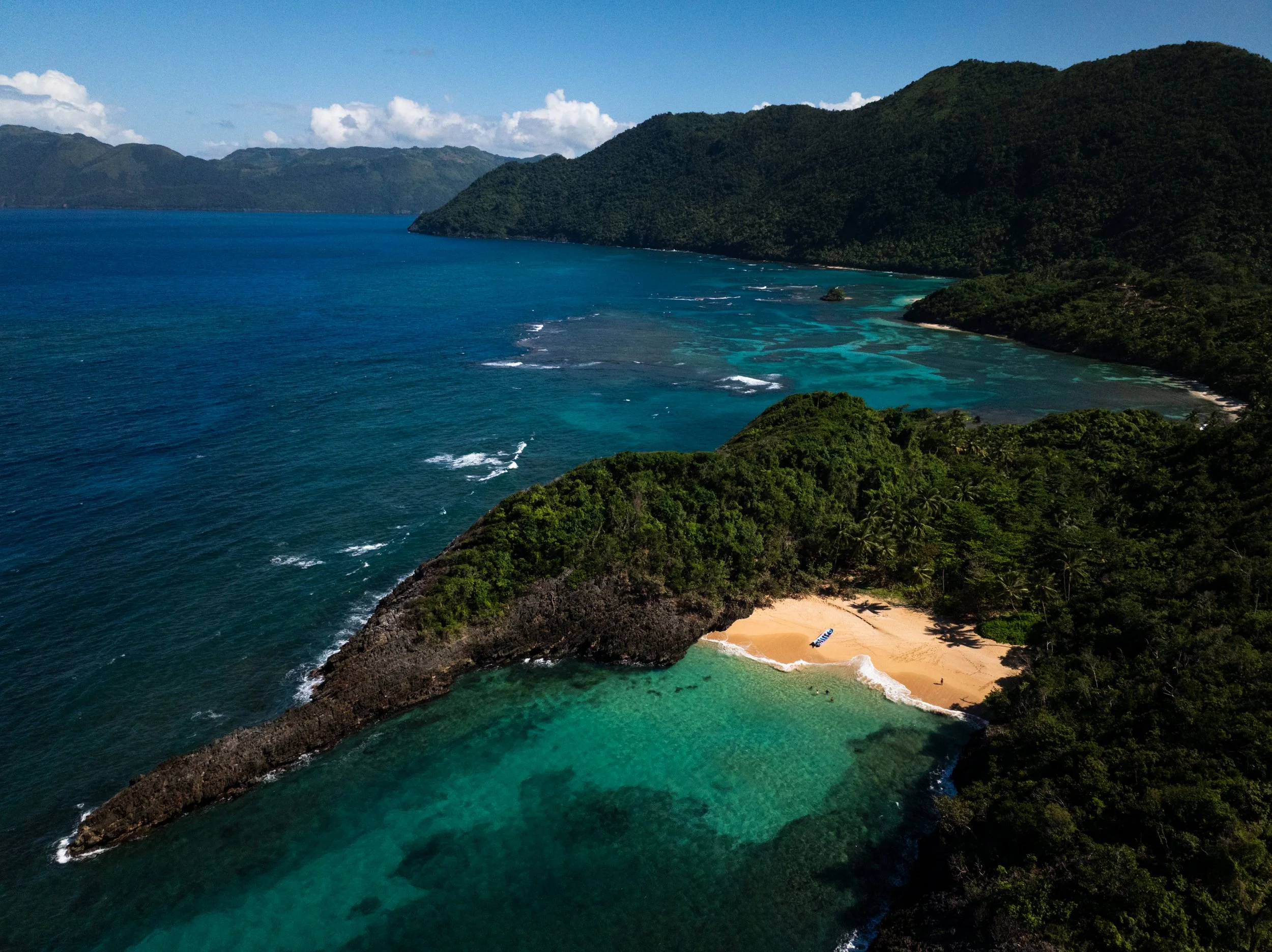 Aerial view of a tropical island with lush green forests, a sandy beach with lounge chairs, and clear turquoise waters.
