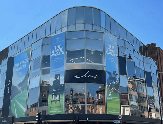 Modern glass building with blue banners announcing 'The Stage Awaits' and posters of athletes, with a black sign displaying the logo 'elys' in the center.