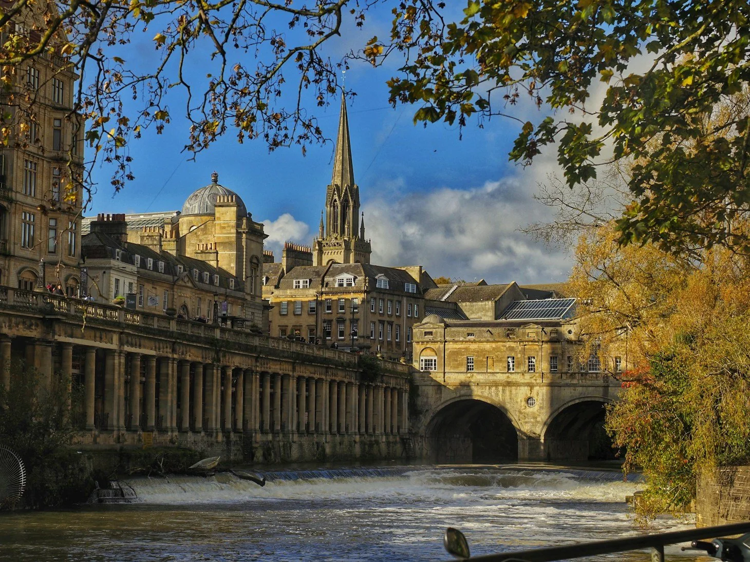 A cityscape featuring historic stone buildings, a church with a tall spire, and a bridge over a river with trees on the riverbank. The sky is blue with some clouds.