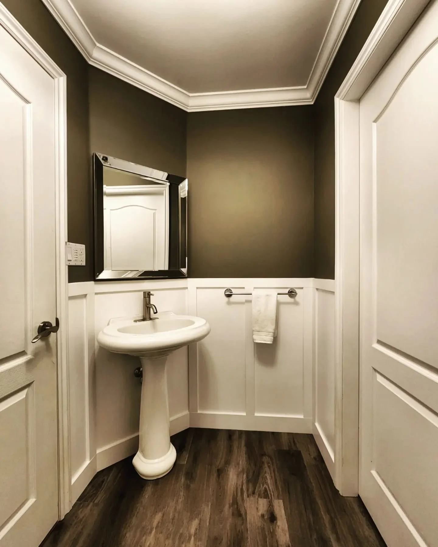 Small powder room with dark gray walls, white wainscoting, a pedestal sink, a rectangular mirror, a towel bar with a towel, and wood-look flooring.