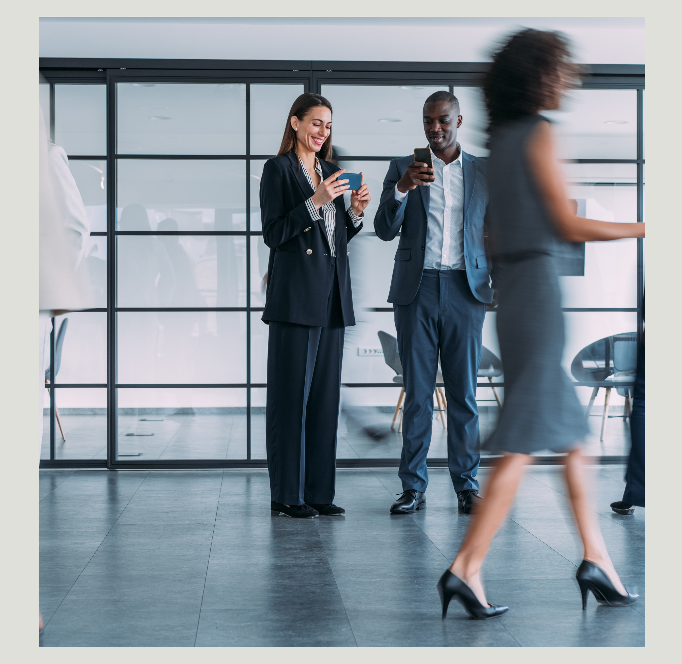 Two professionals, a woman and a man, standing in an office, looking at their smartphones and smiling, while a woman in a gray dress walks past them.
