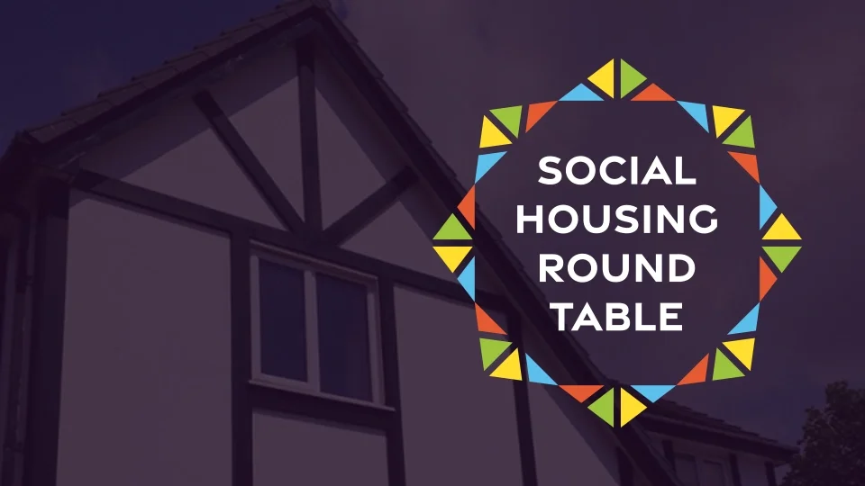 Colorful geometric star-shaped logo composed of multicolored triangles surrounding "Social Housing Round Table" text, overlaid on a residential house background.