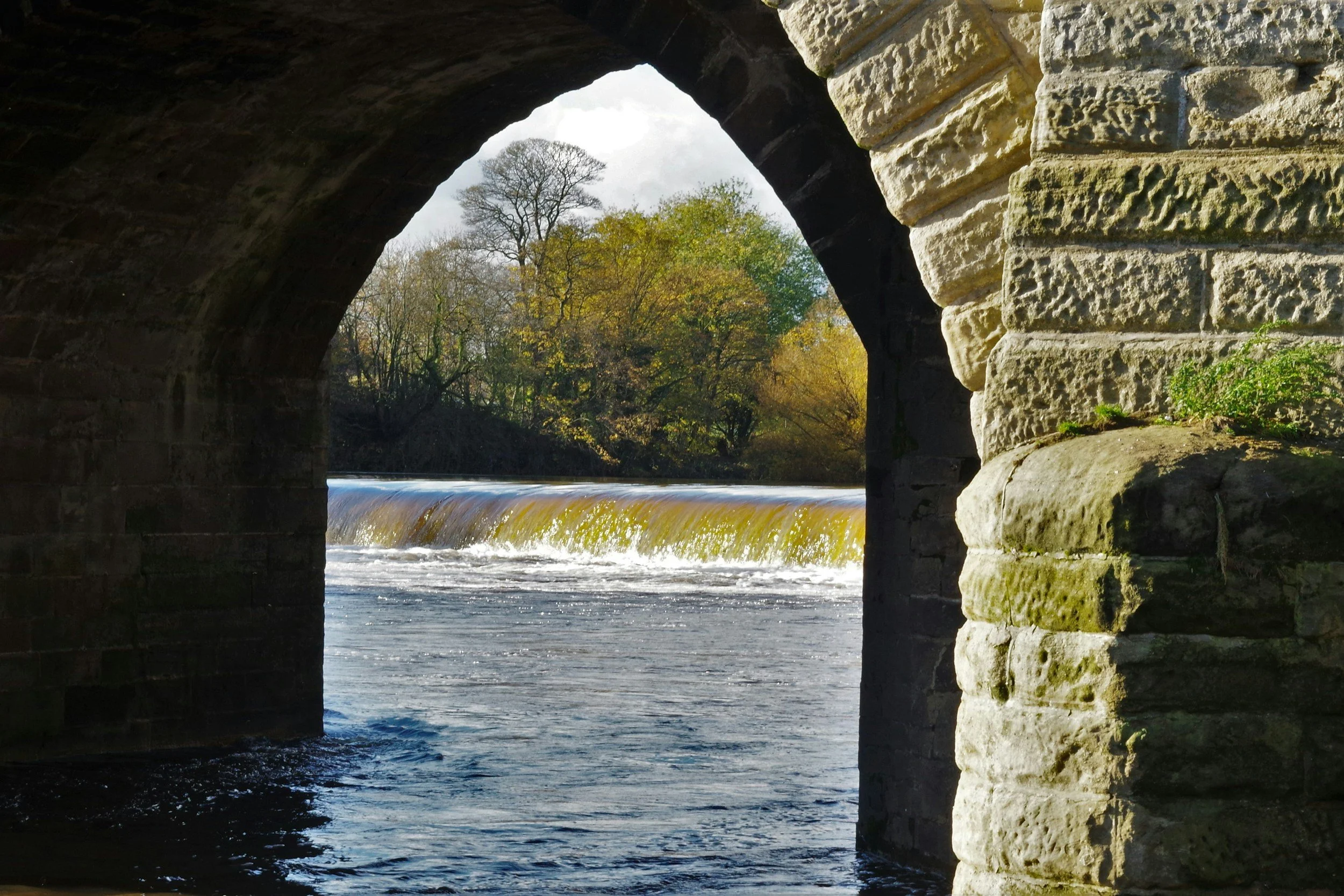 View of a river with a small waterfall or weir seen through an archway of a stone bridge, with trees in the background.