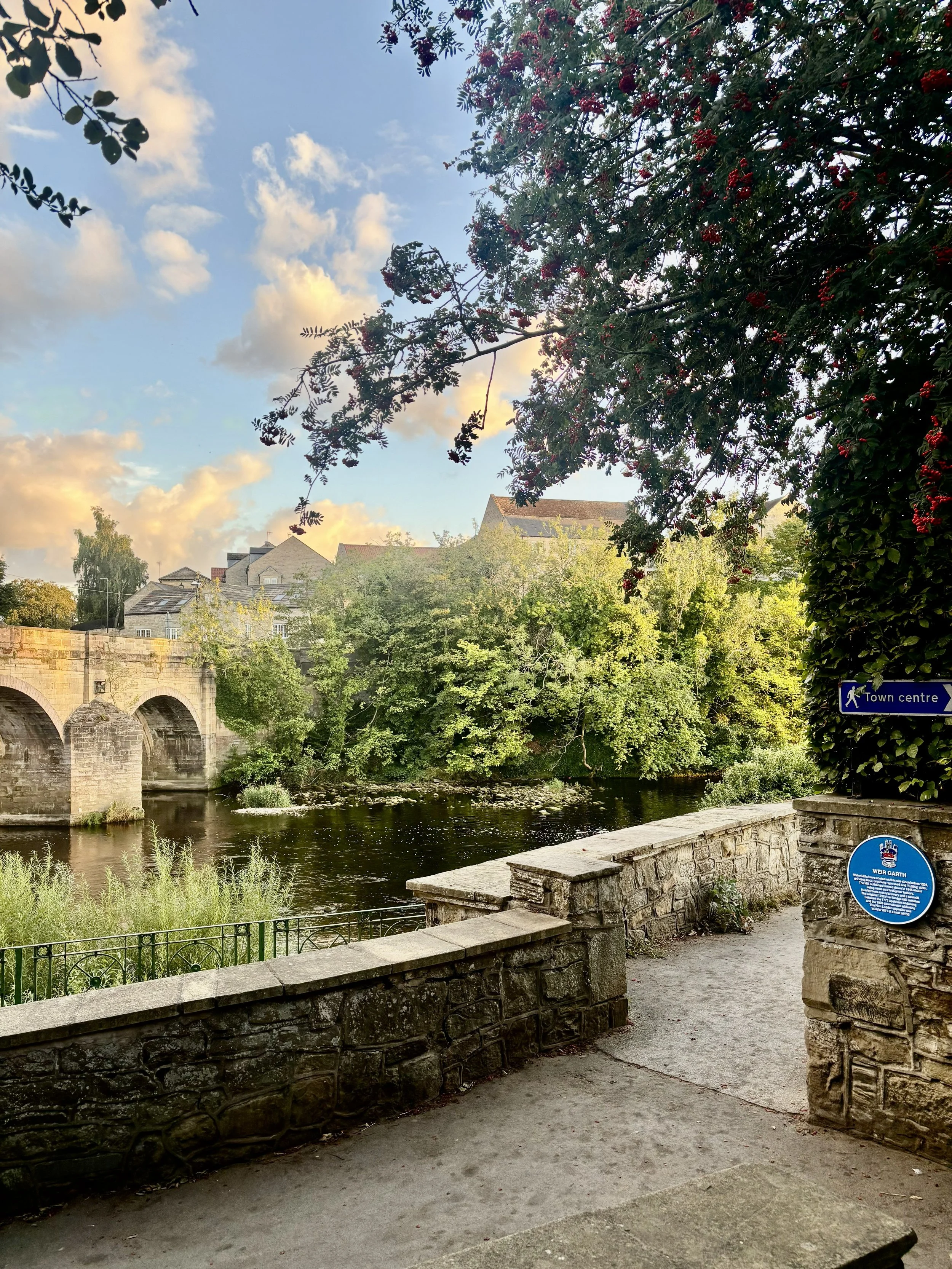 Scenic view of a river with an old stone bridge, trees with green leaves, and a partly cloudy sky. There is a stone path and wall along the riverbank, with signs indicating 'Town centre' and providing historical information.