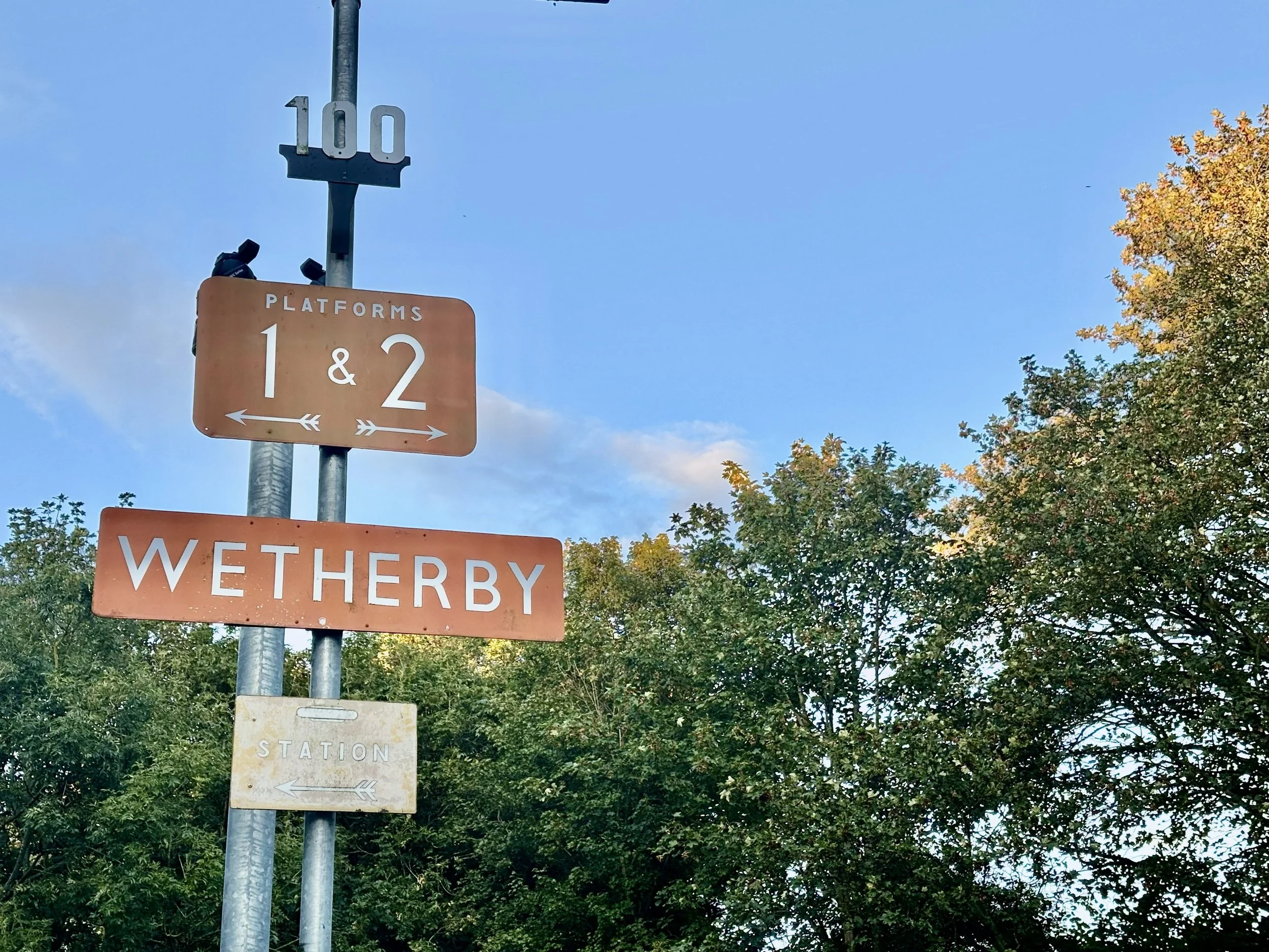 Street signs on a pole at Wetherby station, including platforms 1 & 2 and a former station sign, with trees and a blue sky in the background.