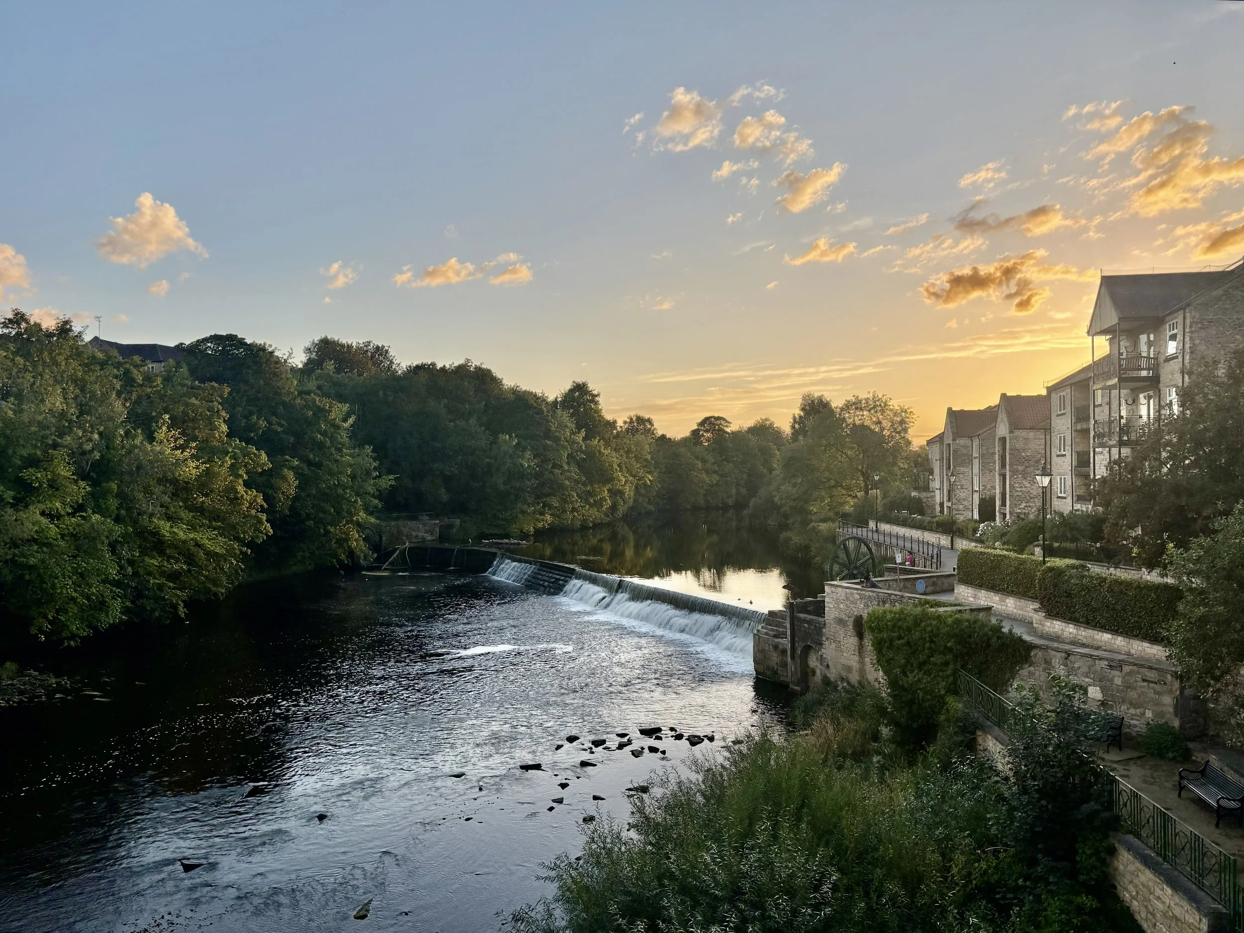 A serene riverside scene at sunset with a small waterfall, lush green trees on both sides, and residential buildings on the right with balconies and outdoor seating.