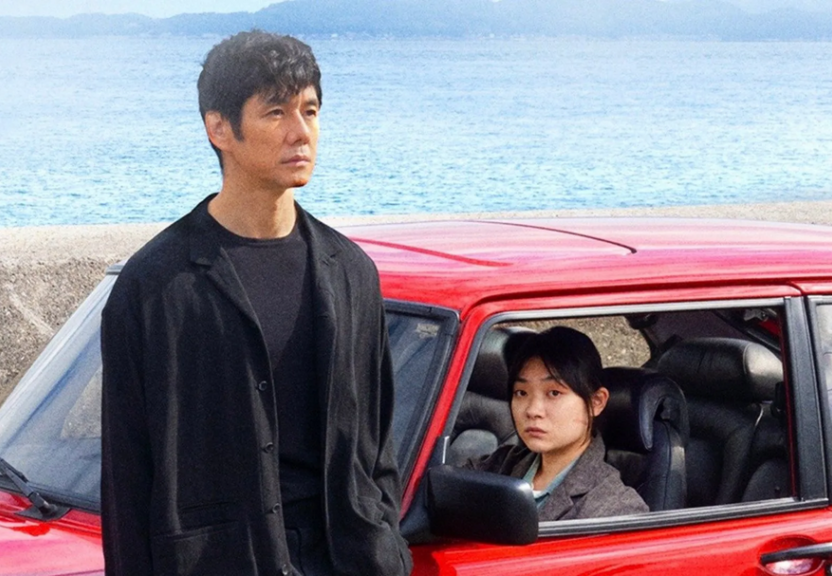 A man standing outside a red car with a woman sitting inside near a body of water.