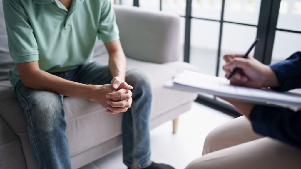 Person wearing a green polo shirt and jeans sitting on a sofa during a therapy or counseling session with a counselor taking notes.