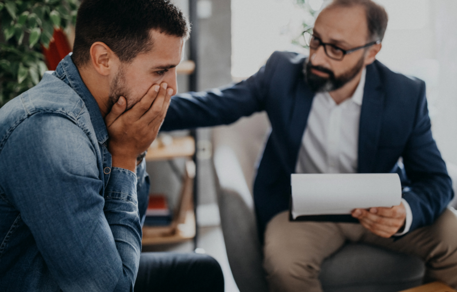 A man in a denim shirt appears distressed, covering his mouth with his hands, as a therapist or counselor in a suit and glasses gently places a hand on his shoulder during a counseling session.