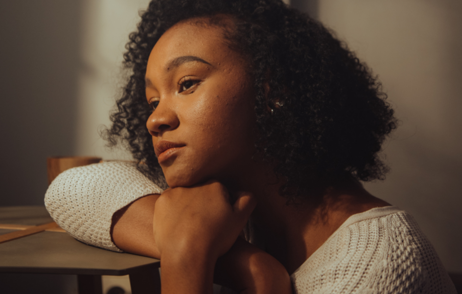 A young woman with curly hair resting her chin on her arm on a desk, looking contemplative.