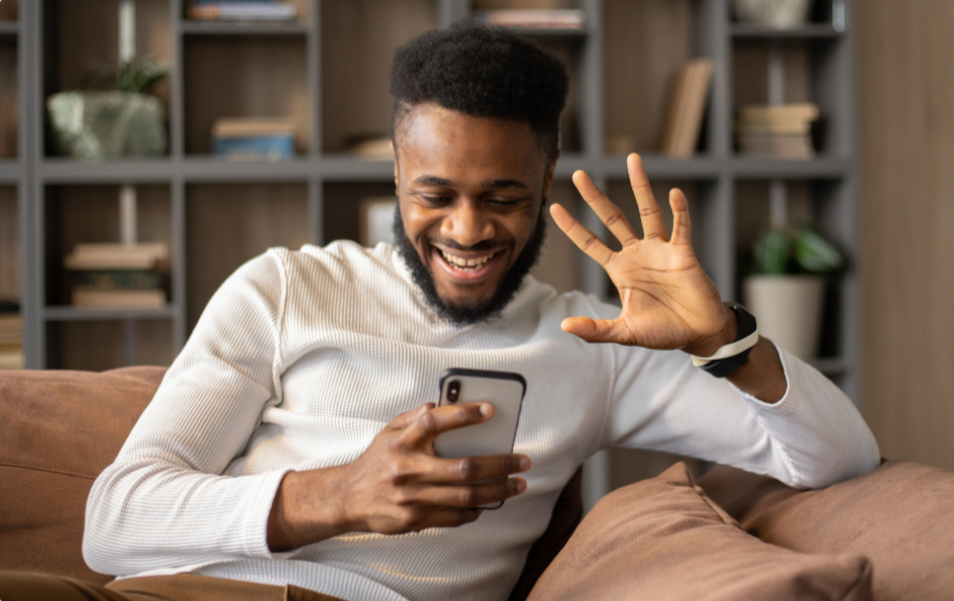 A young man with curly hair and a beard sitting on a sofa, smiling, looking at his phone, waving with his right hand, in a cozy living room with a bookshelf in the background.