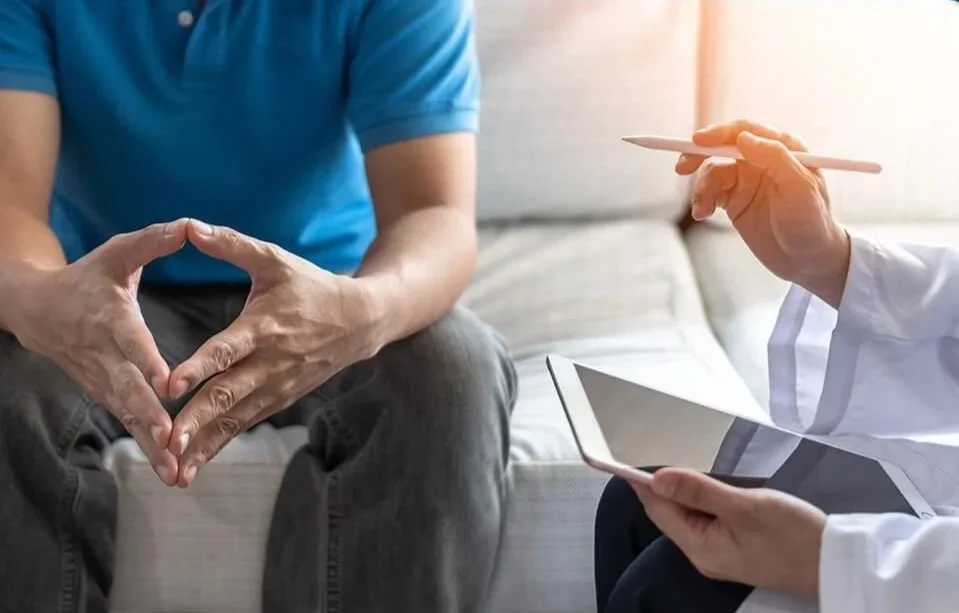 Patient sitting with hands clasped in a triangular shape during a consultation with a healthcare professional, who is holding a tablet and a pen