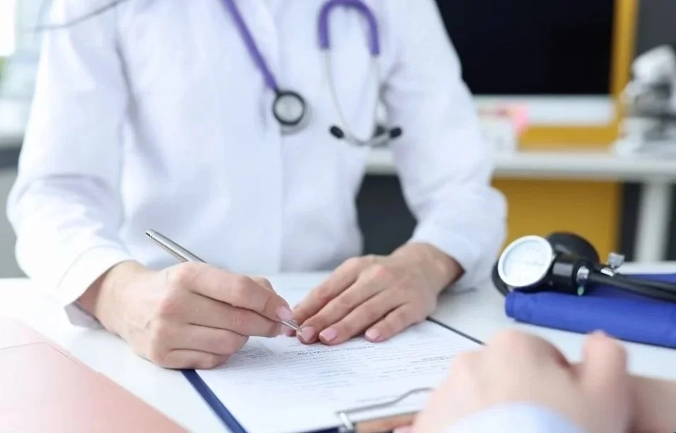 A healthcare professional wearing a white coat and stethoscope writing on a document at a desk.