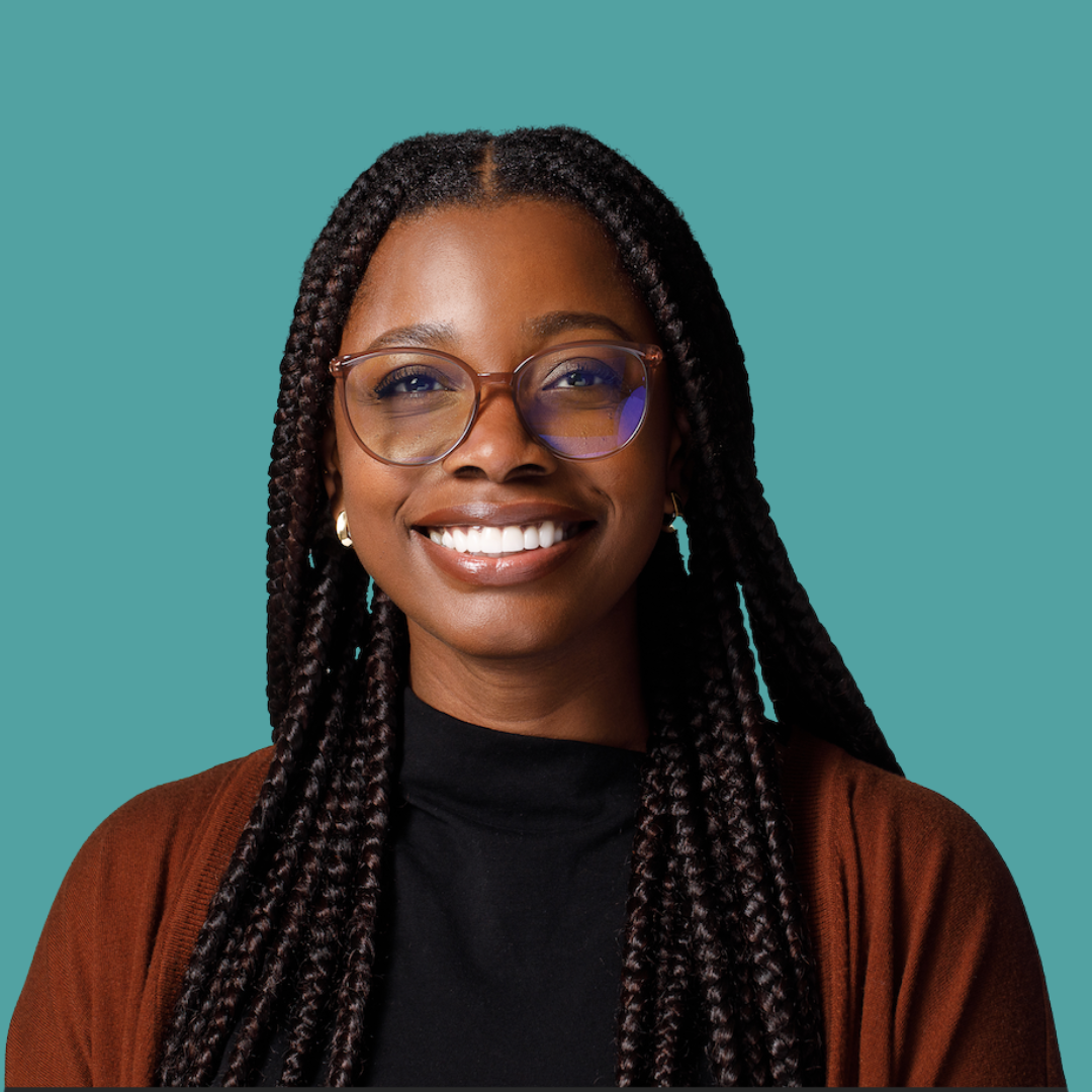 A woman with long braided hair, wearing glasses and a brown cardigan, smiling in front of a teal background.