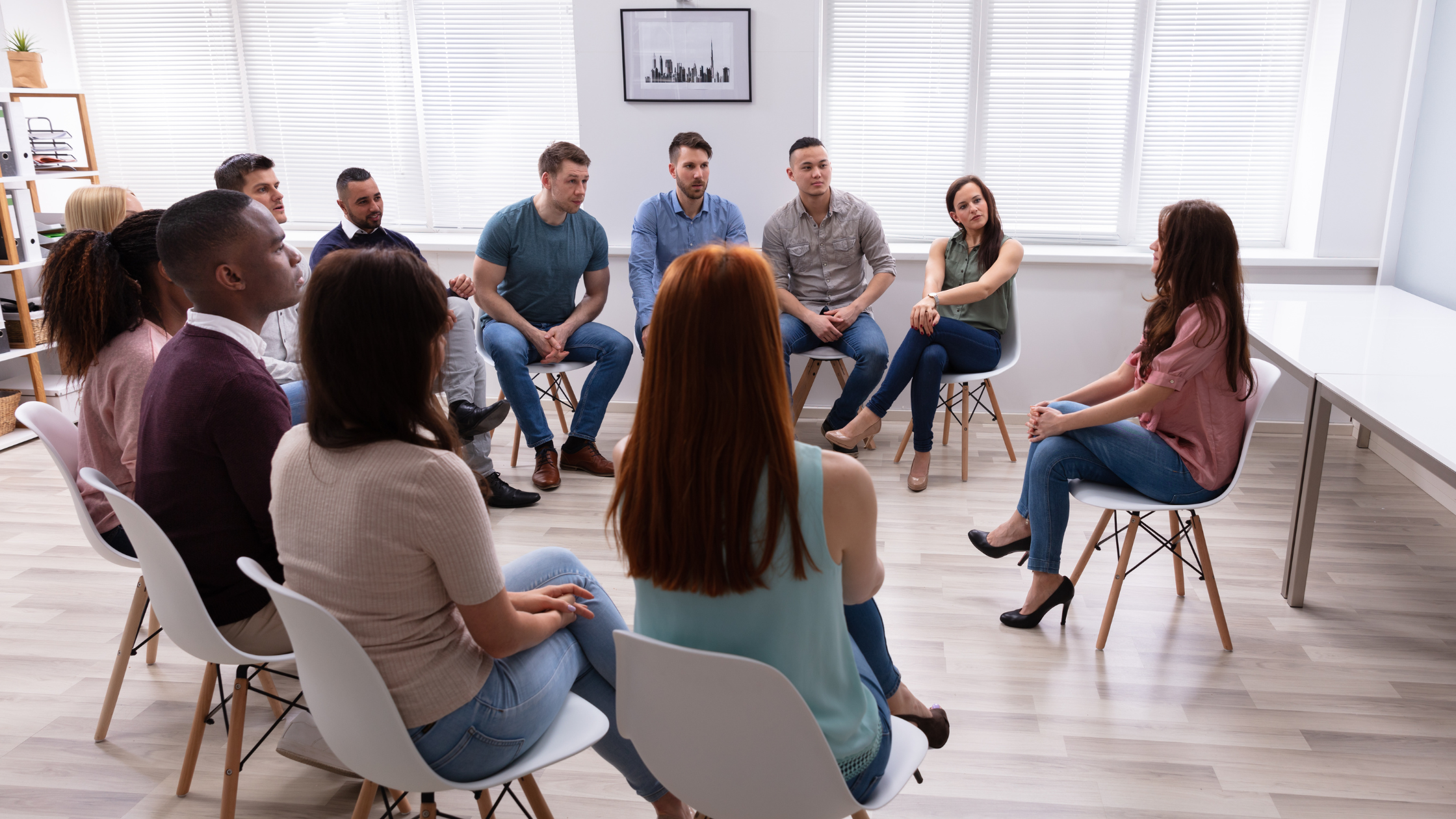 A diverse group of people participating in a discussion in a bright, modern office with large windows and white blinds.