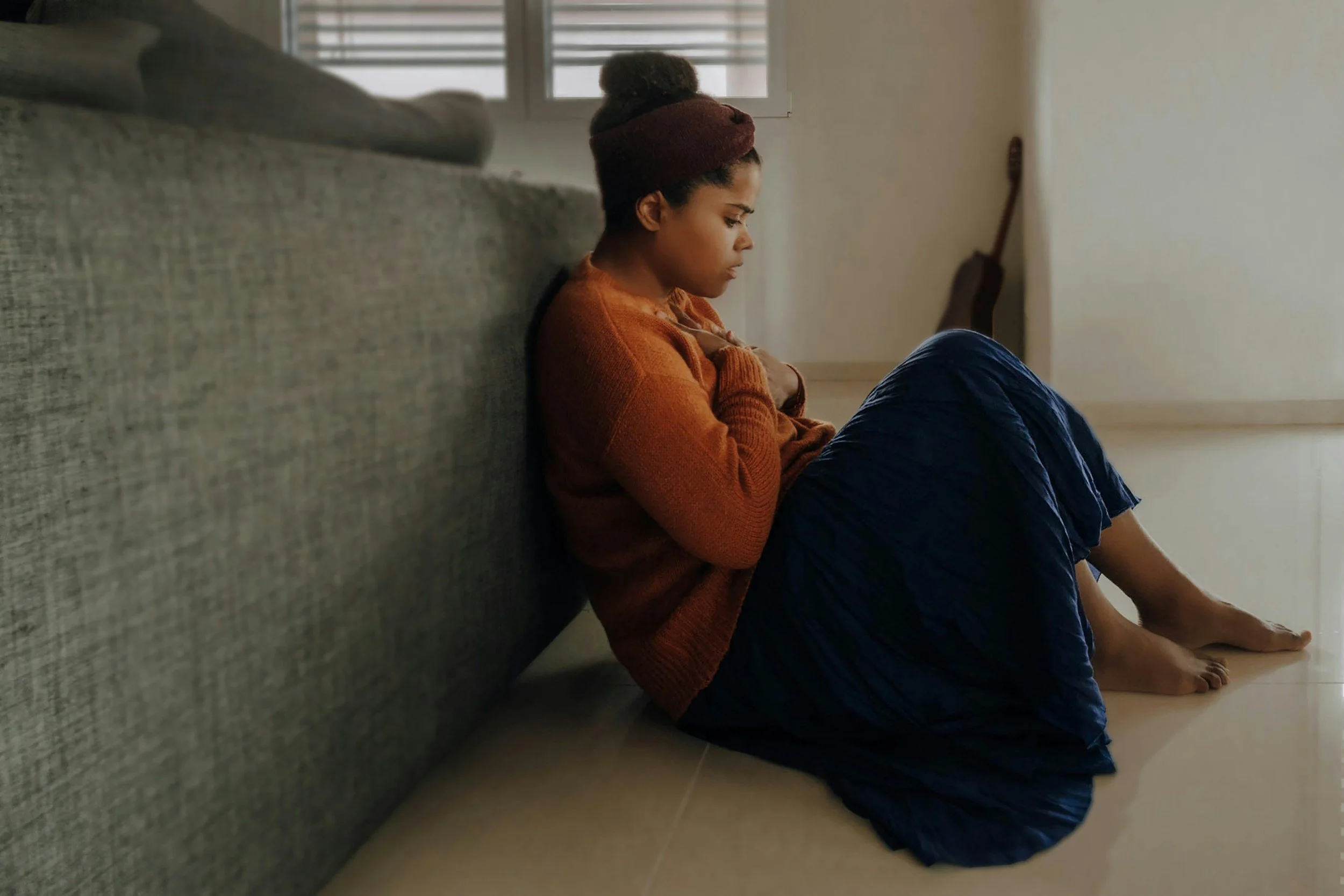 A woman sitting on the floor with her back against a sofa, wearing a brown headband, an orange sweater, and a dark blue skirt, looking distressed with her hands on her chest in a minimalist room with a guitar in the background.