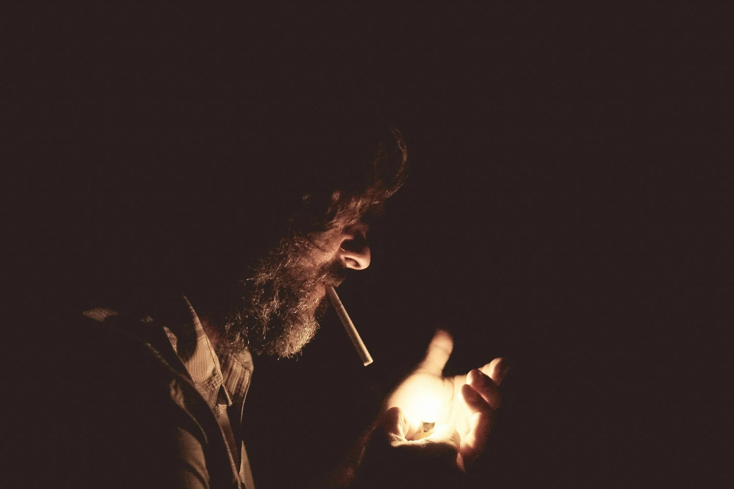 A man with a beard smoking a cigarette at night, looking at a glowing object in his hand.