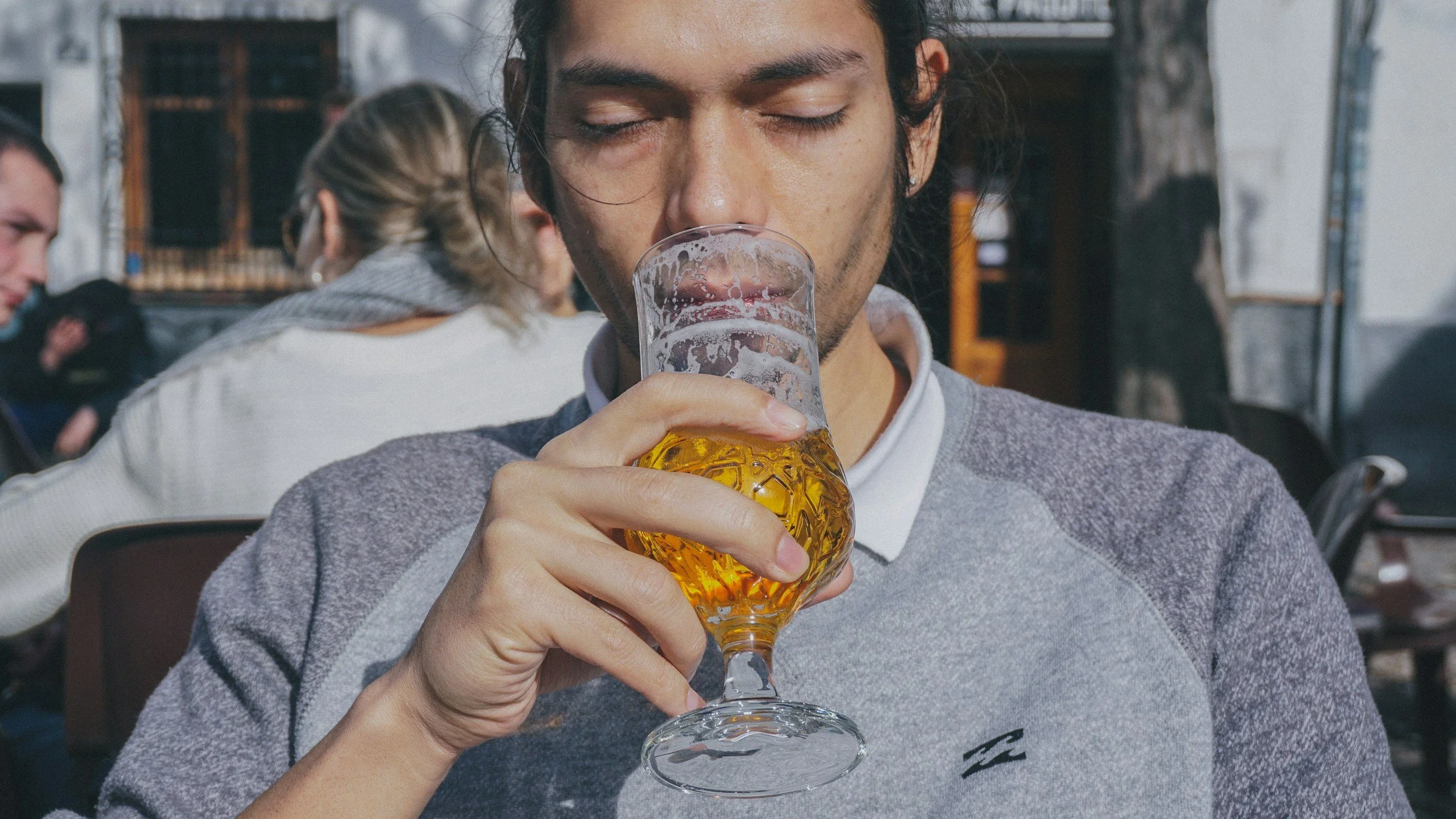A young man with dark hair and closed eyes is drinking a glass of beer outside at a social gathering.