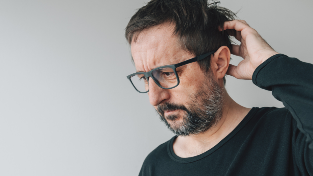 A man with glasses and a beard wearing a black long sleeve shirt, touching the back of his head, looking down with a pensive expression.