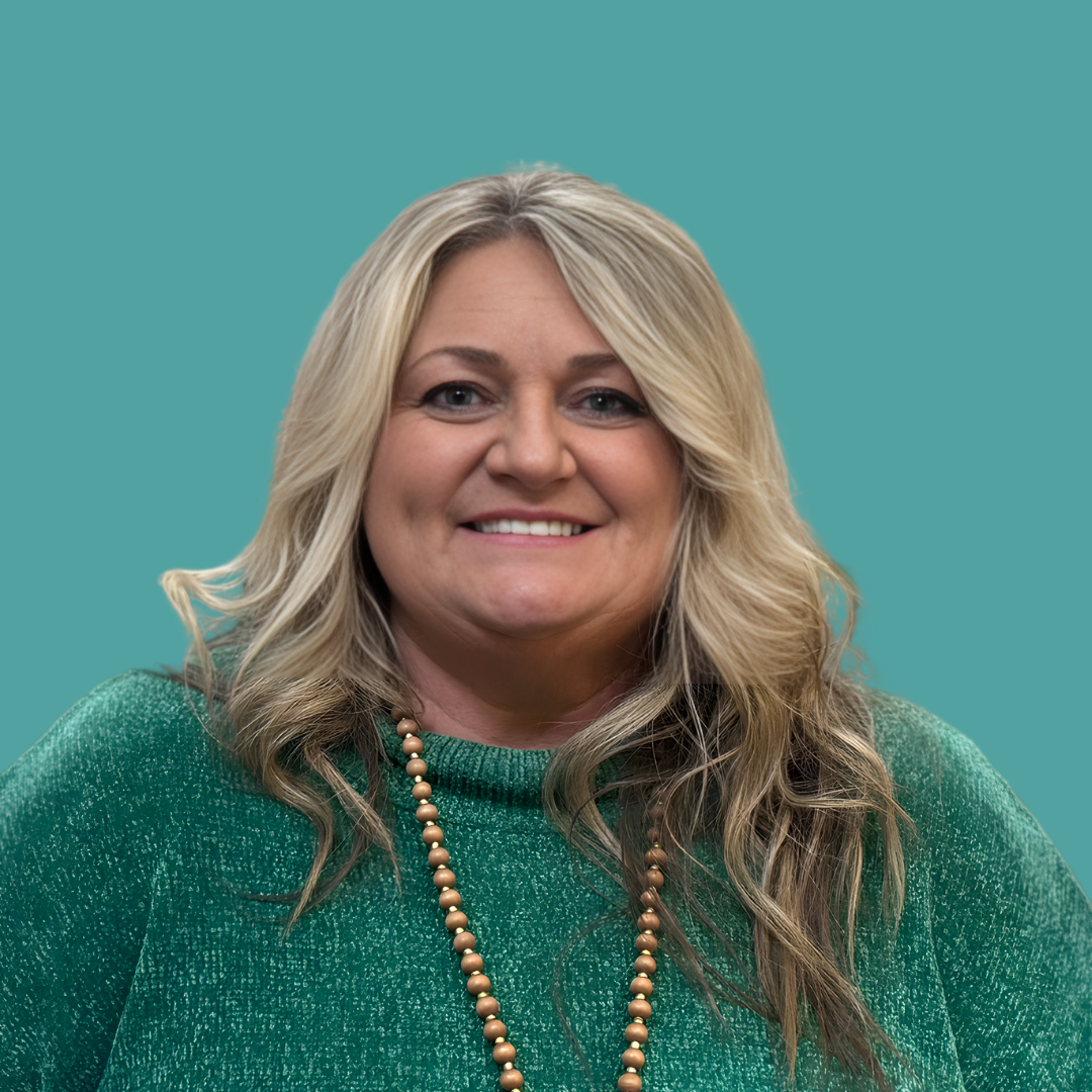 A woman with blonde hair smiling, wearing a green sweater and a beaded necklace, standing against a teal background.