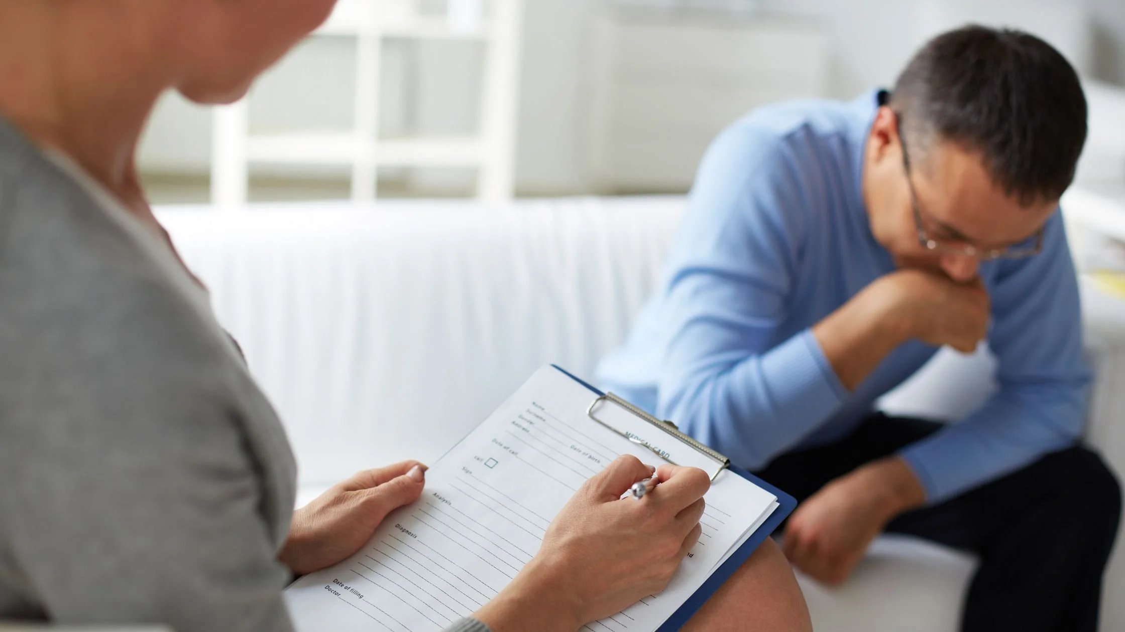 A woman filling out a medical form on a clipboard while a man in a blue shirt sits on a couch, leaning forward and covering his mouth with his hand, possibly coughing or thinking, in a waiting room.