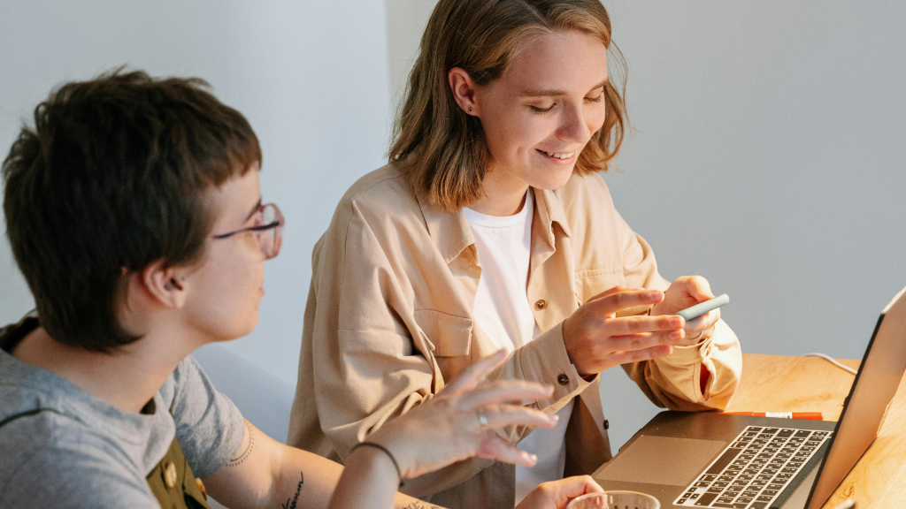 Two women sitting at a wooden table, one with glasses, looking at the other who is smiling and holding a smartphone. There is a laptop on the table.