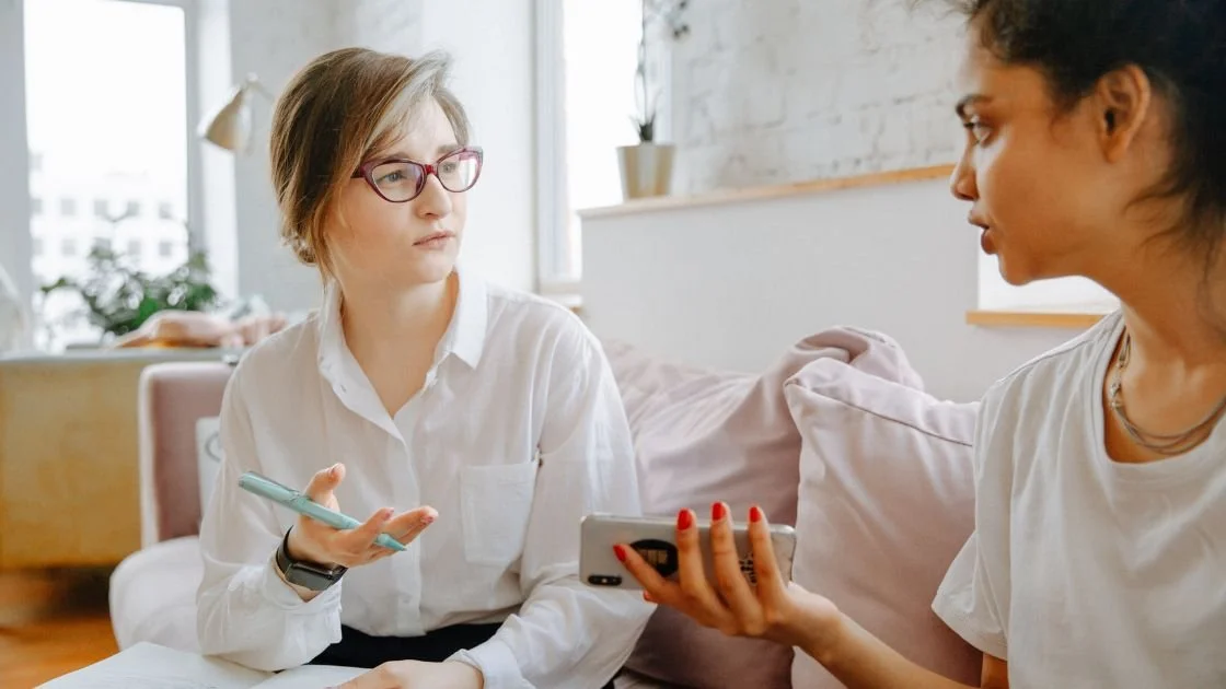 Two women having a serious conversation in a well-lit room, one holding a phone and the other holding a pen.