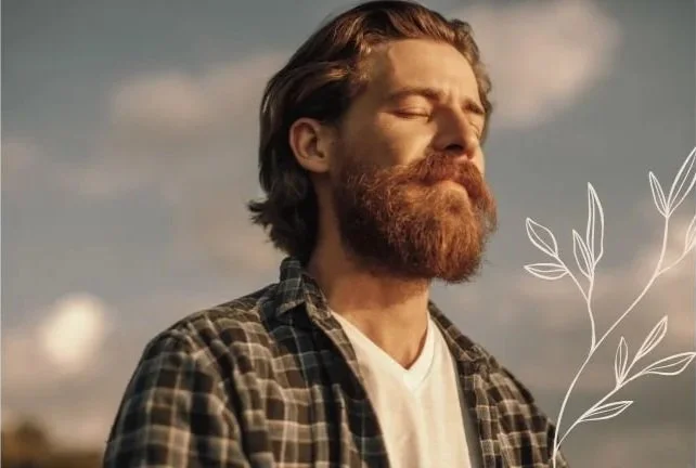 A man with a beard and closed eyes is outdoors on a sunny day, wearing a plaid shirt over a white T-shirt.