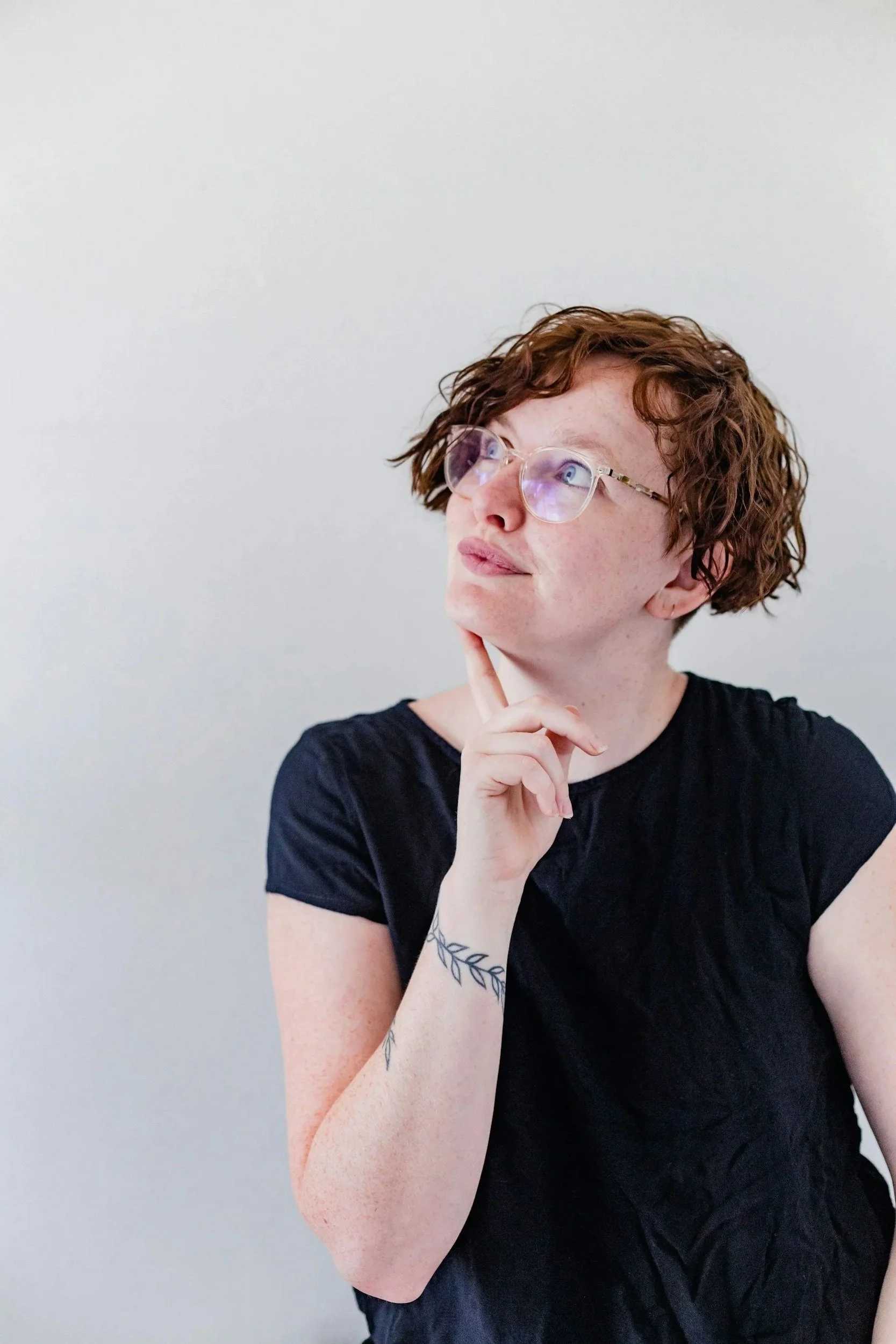 A person with short, curly red hair and glasses, wearing a black shirt, is looking thoughtful with their chin resting on their hand, against a plain light background.