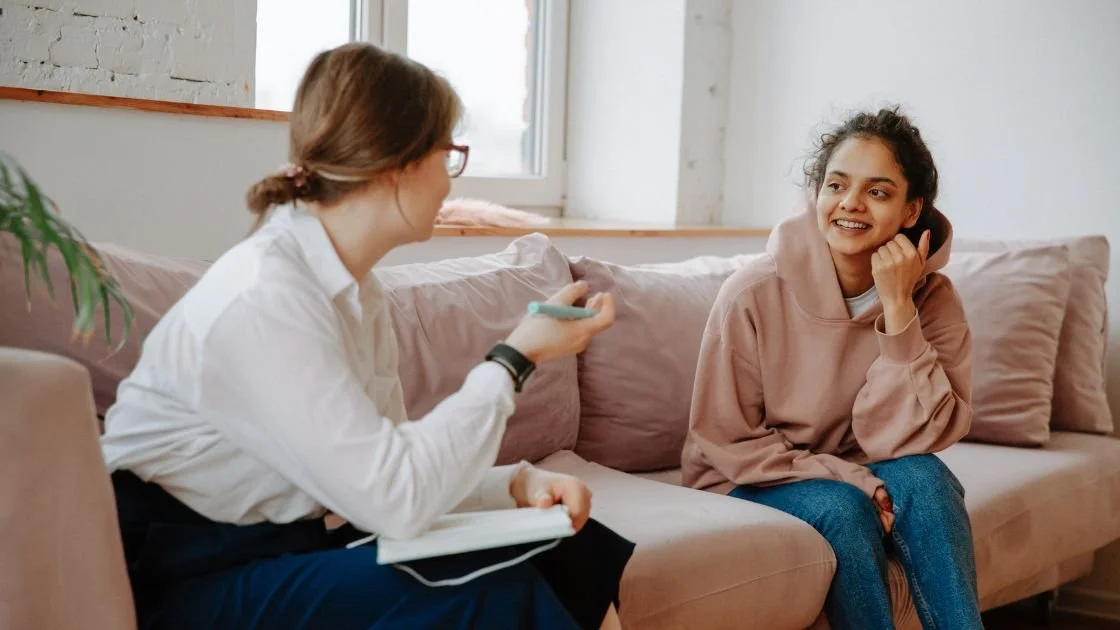 A woman in a white shirt and glasses sitting on a sofa during a therapy session or counseling, talking to a young woman in a pink hoodie and blue jeans, who is smiling and holding a phone to her ear.