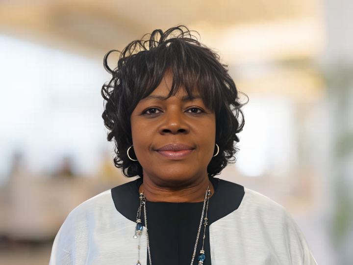 A woman with short, curly black hair wearing a black and white top and a necklace, standing indoors with a blurred background.